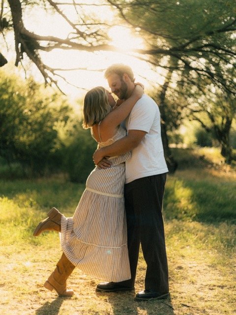 A couple hugging and smiling in a sunlit outdoor setting with trees in the background.