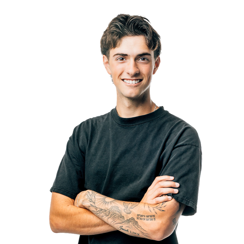 A young man with short brown hair, smiling, wearing a black t-shirt and has tattoos on his left arm, standing with arms crossed.