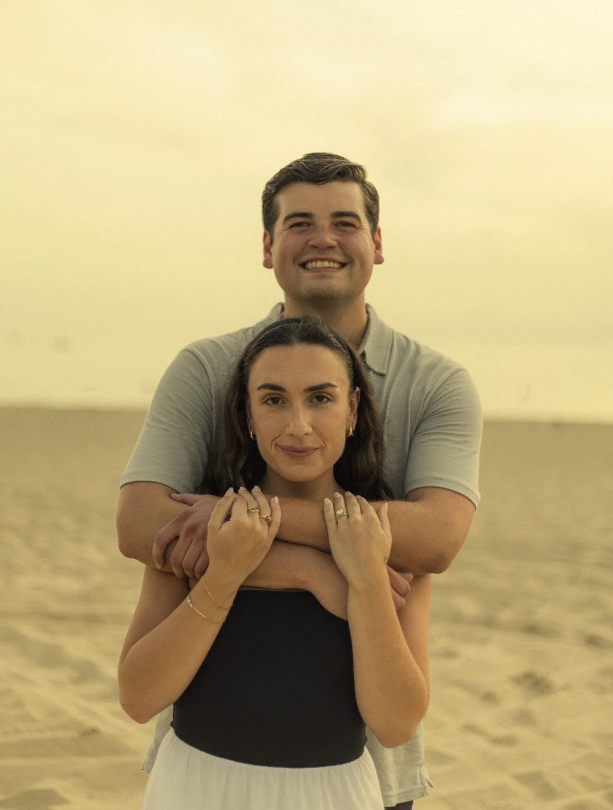 A smiling man and woman standing on a beach. The man is behind the woman with his arms around her shoulders. The woman is facing forward with her hands resting on top of the man's arms. The background shows the beach and ocean with a yellowish sky.