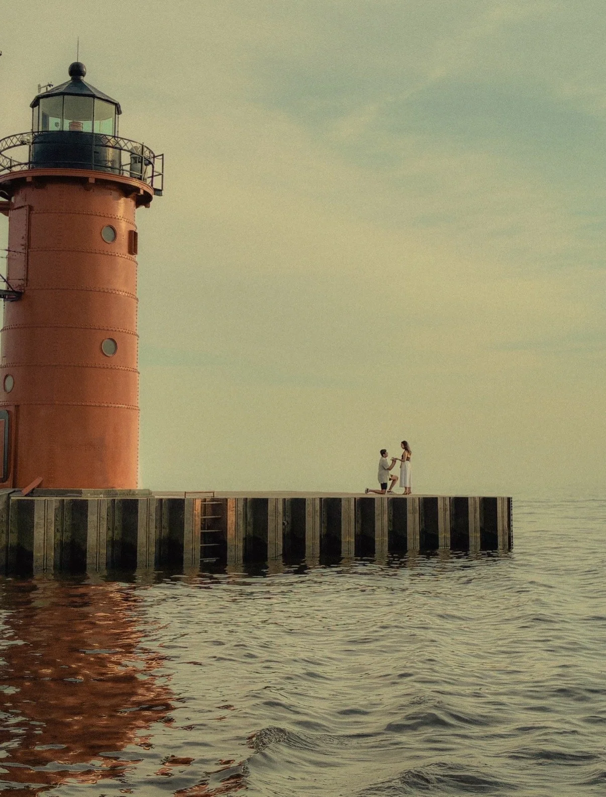 A man kneeling on one knee, holding a woman's hands while proposing, on a dock next to a large red lighthouse by the water during sunset.