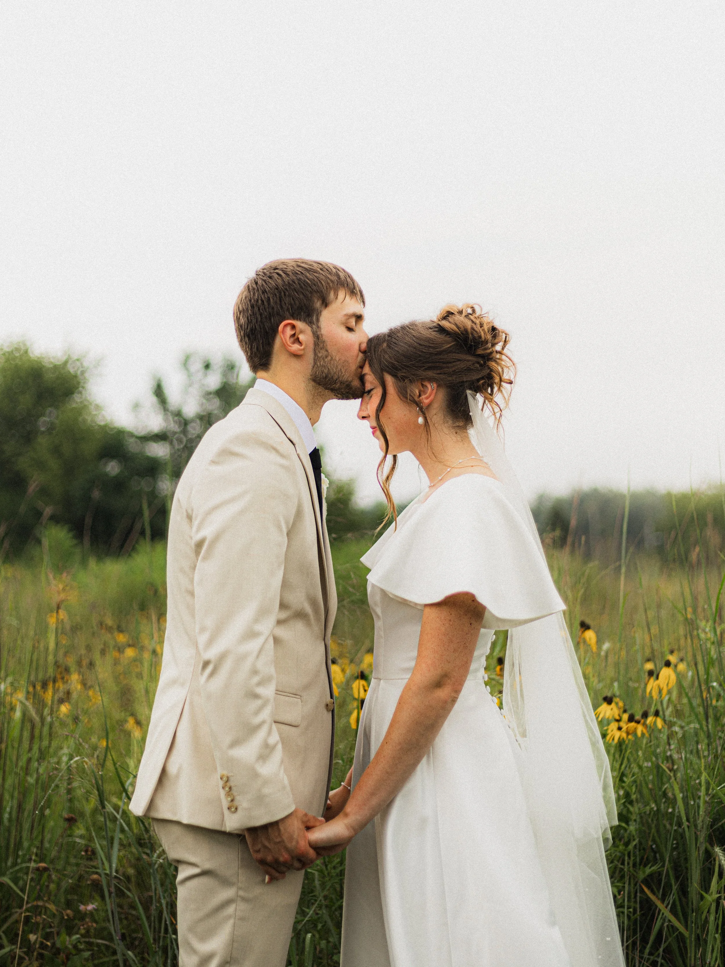A bride and groom holding hands with foreheads touching, standing outdoors in a grassy field with yellow flowers, during their wedding ceremony.