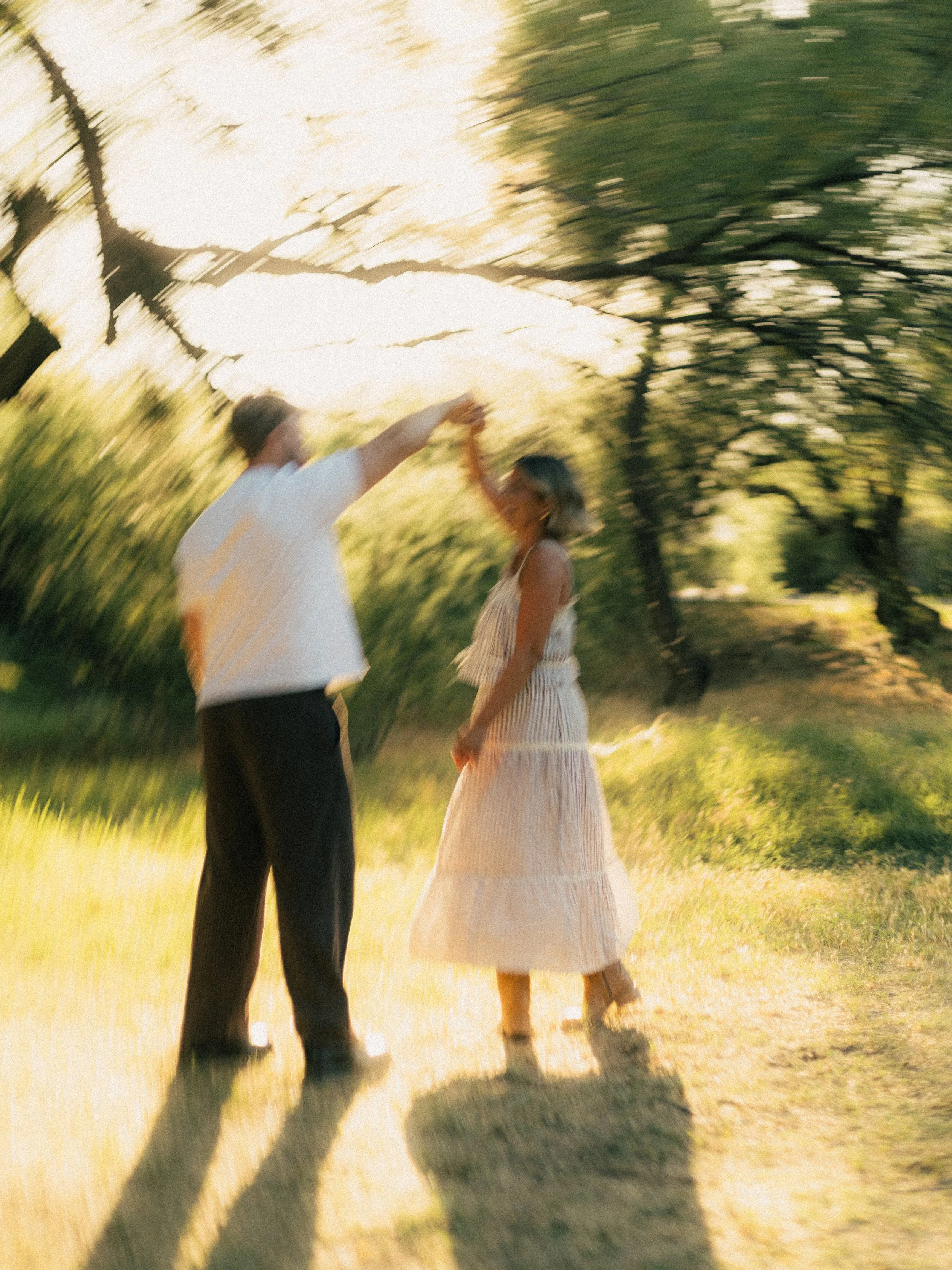 A blurred photo of a couple holding hands, dancing or walking outdoors in a natural setting with trees and grass, bathed in warm sunlight.