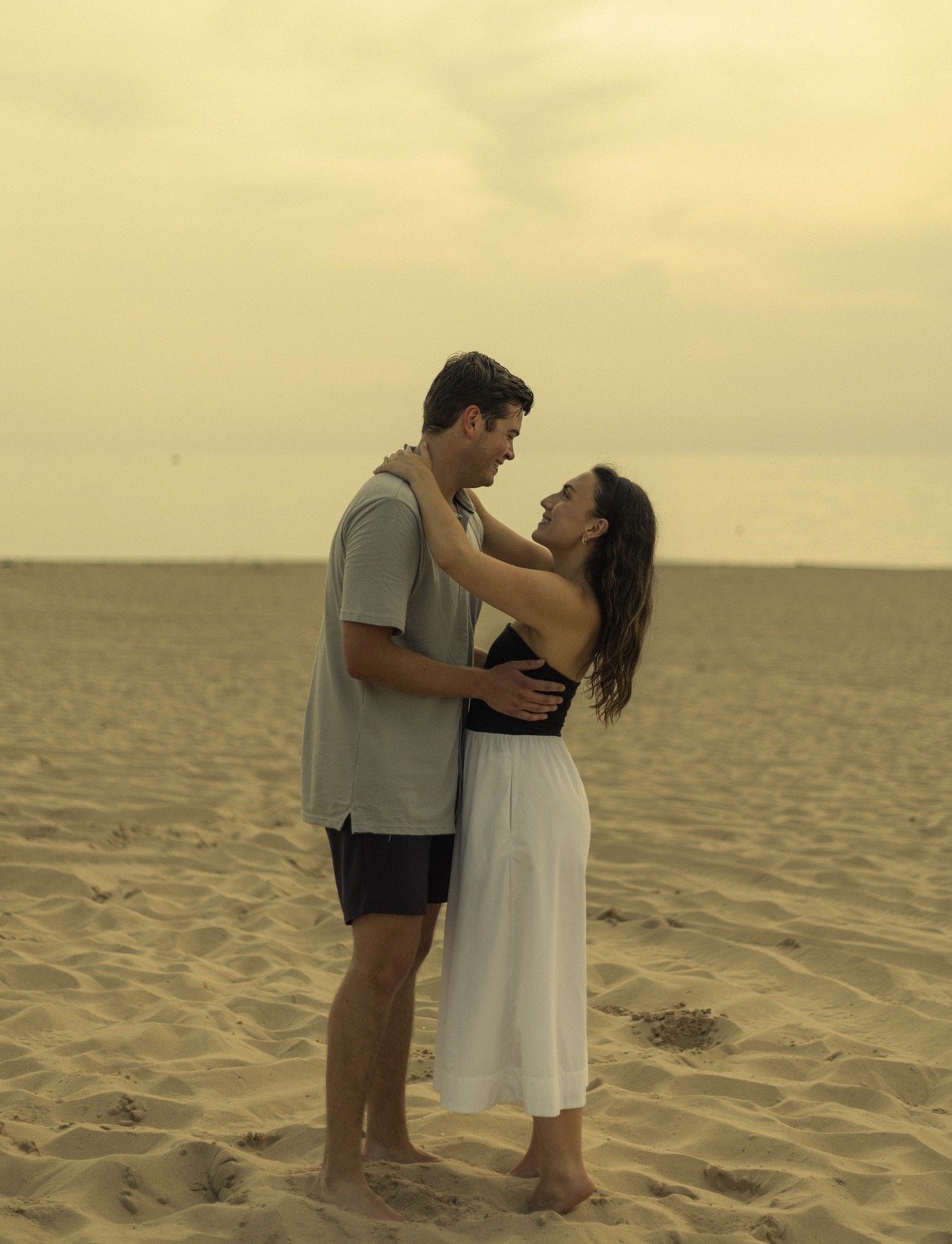 A young couple standing close together on a sandy beach at sunset, looking into each other's eyes and smiling, with the ocean in the background.
