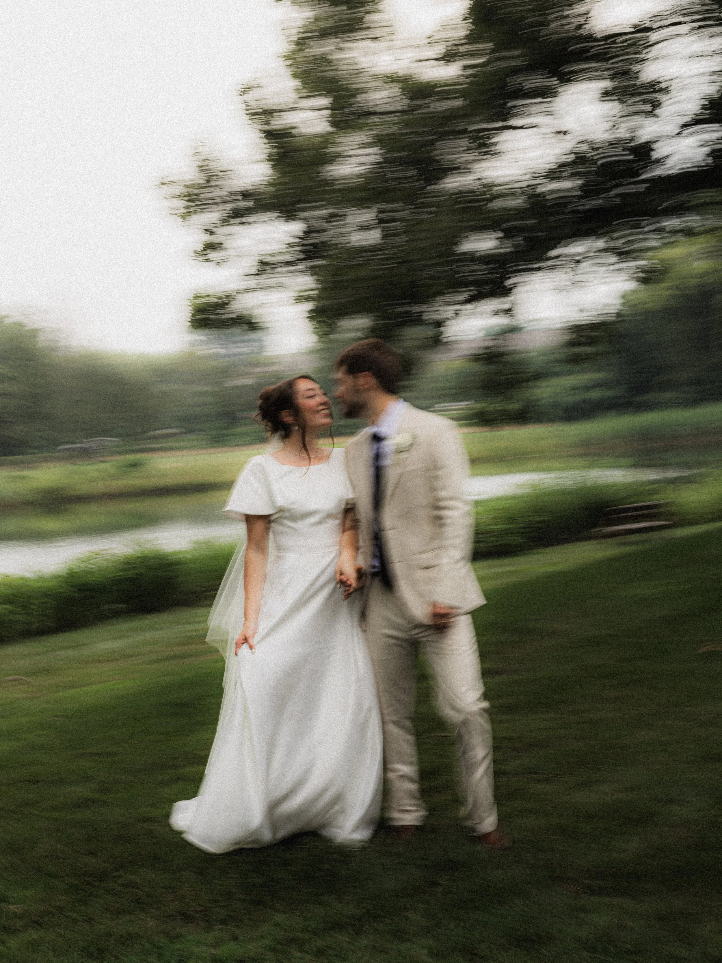 A bride and groom standing outdoors, holding hands, with a blurred background of trees and grass.