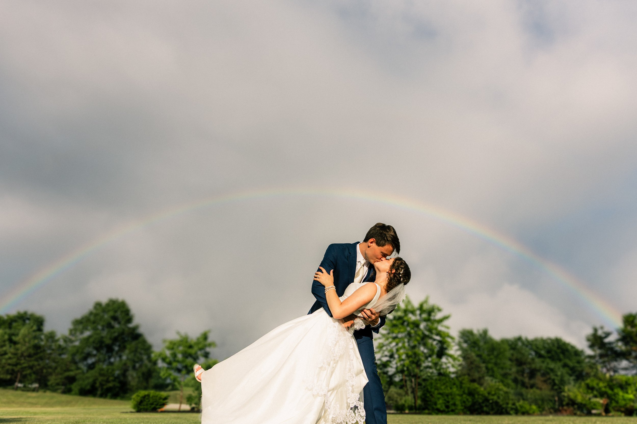 A wedding couple sharing a kiss outdoors under a rainbow, with lush green trees and a cloudy sky in the background.