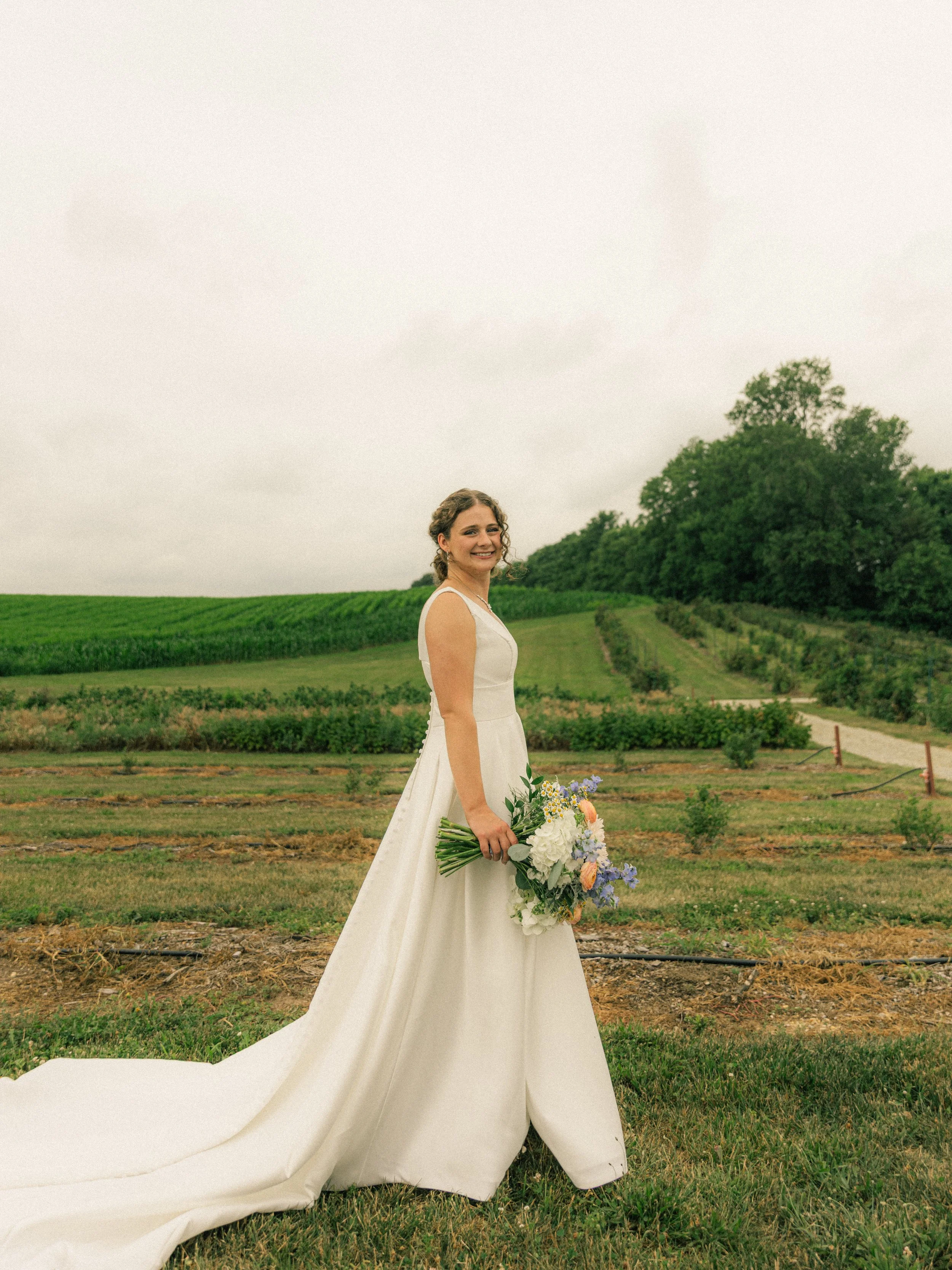 A bride in a white wedding gown holding a bouquet of flowers, standing in a lush green field with rows of plants, trees in the background, and a cloudy sky above.