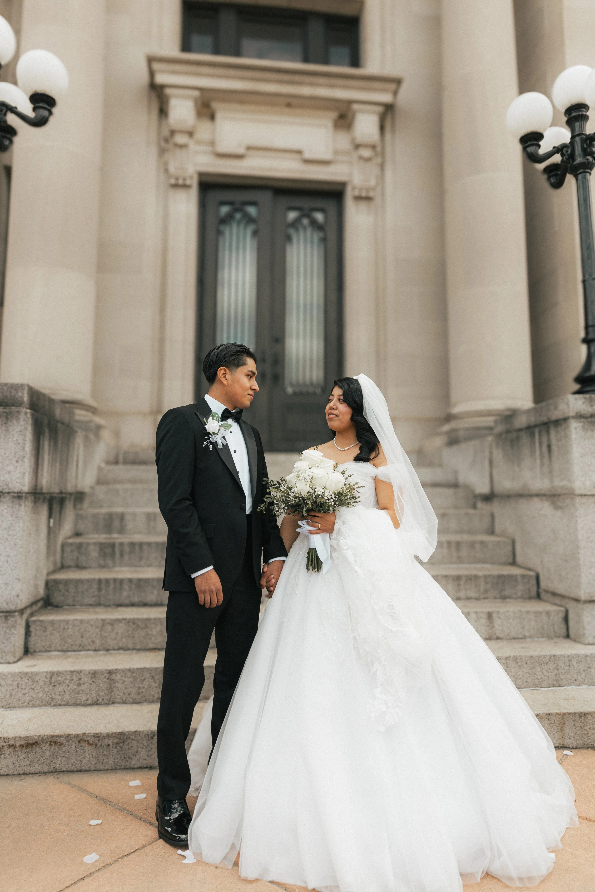 A bride and groom standing on steps outside a building, holding hands and looking at each other, dressed in wedding attire. The bride is wearing a white wedding gown and veil, while the groom is in a black tuxedo with a boutonniere.