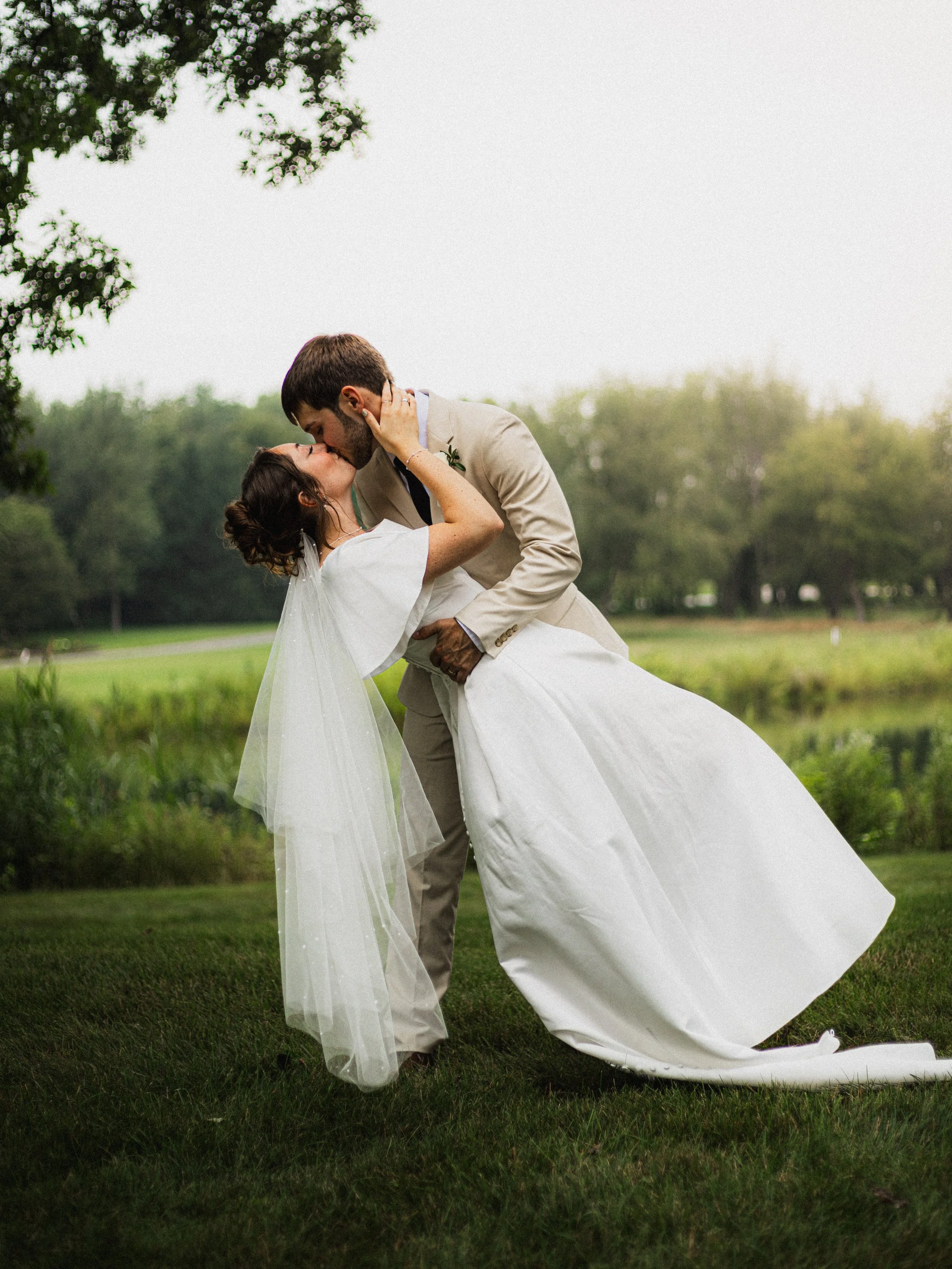 A bride and groom kissing outdoors on a grassy field with trees in the background, the groom dipping the bride and holding her waist.