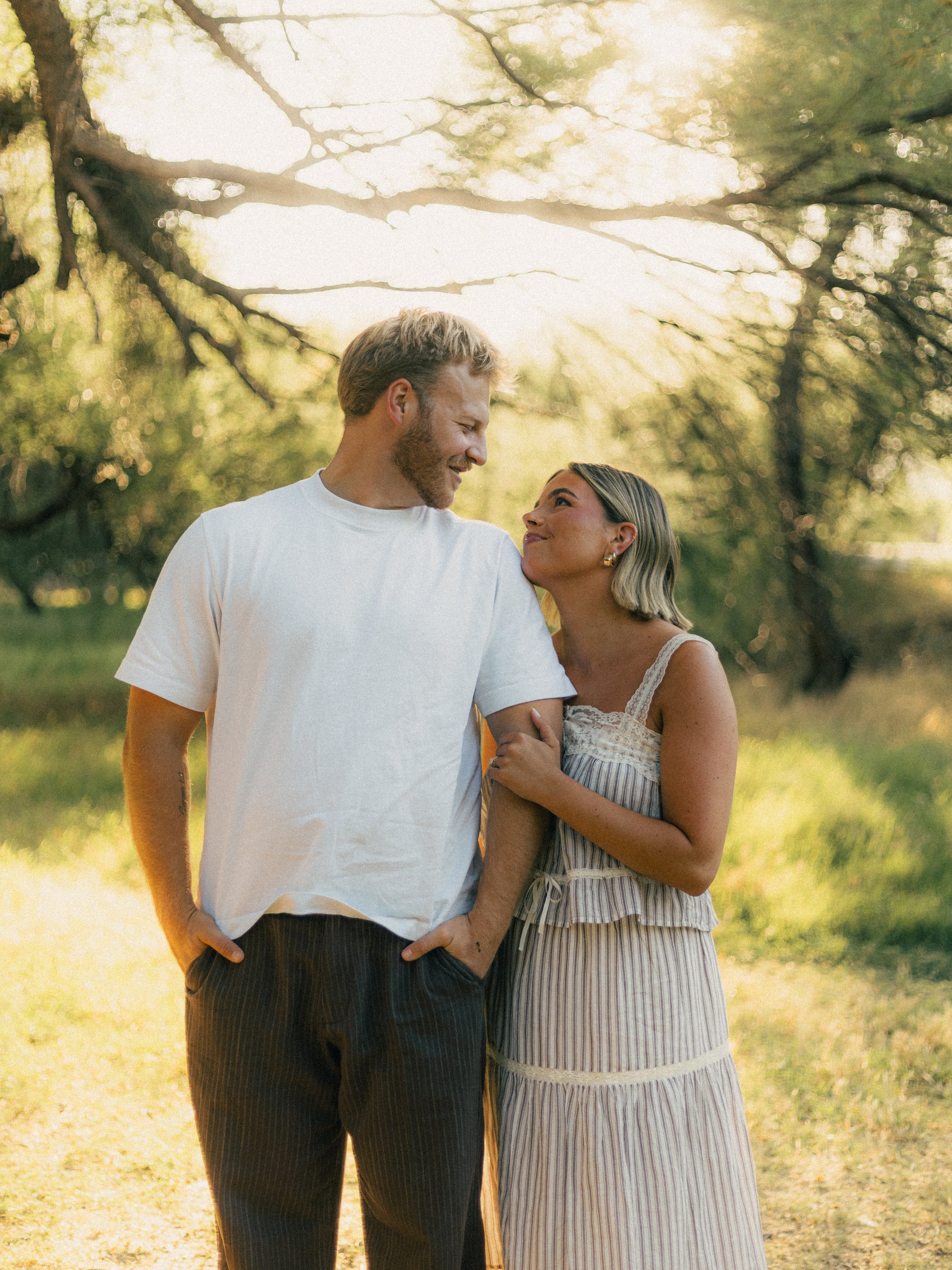A couple standing close together outdoors, enjoying a sunny day in a park with trees in the background. The man is wearing a white t-shirt and striped pants, smiling down at the woman, who is looking up at him with a smile, wearing a striped dress an