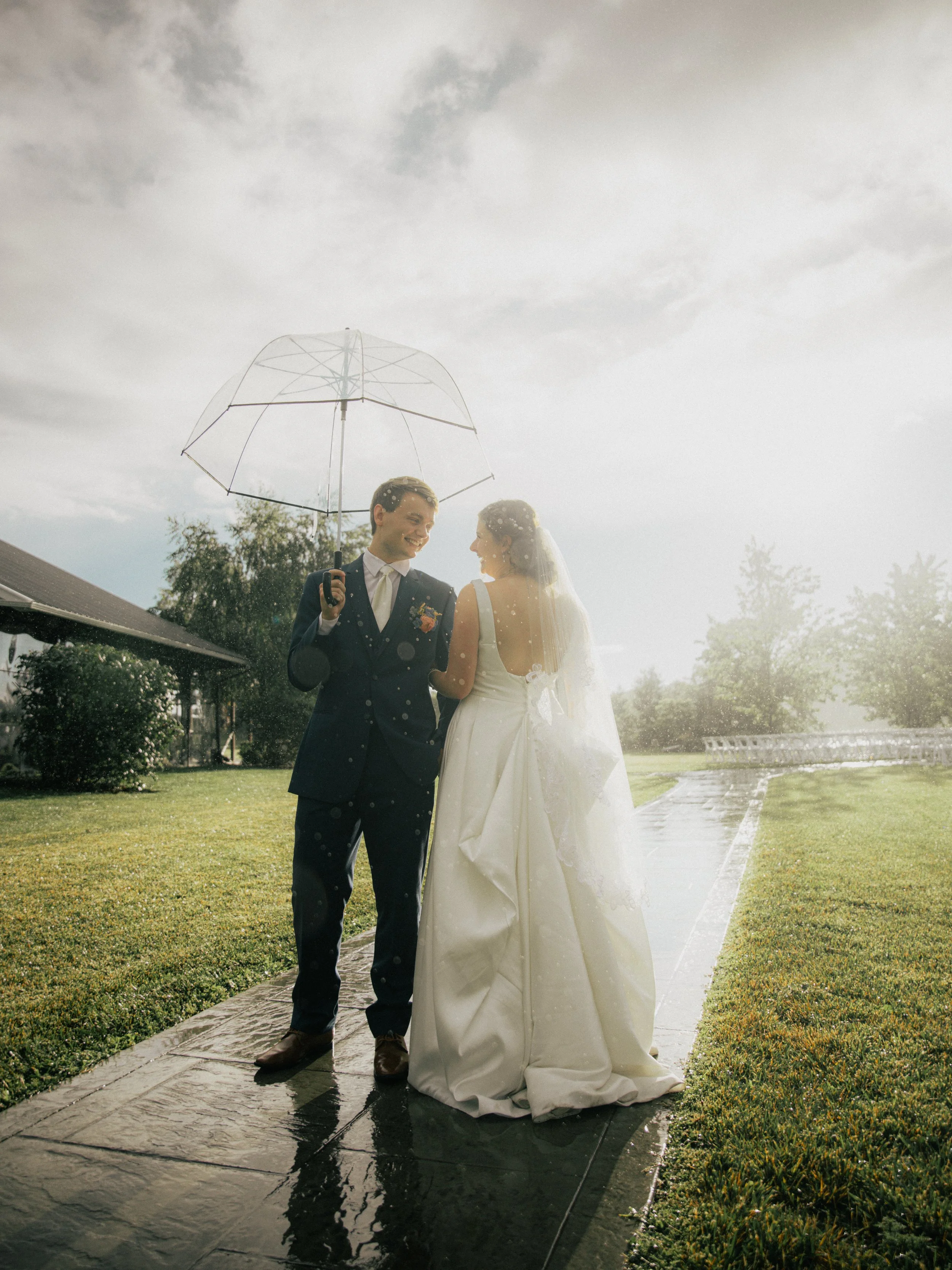 A newlywed couple standing on a wet sidewalk outside during a rain shower, sharing a joyful moment under an umbrella. The bride is wearing a white wedding dress with a veil, and the groom is in a dark suit with a tie and boutonniere. The sky is cloud