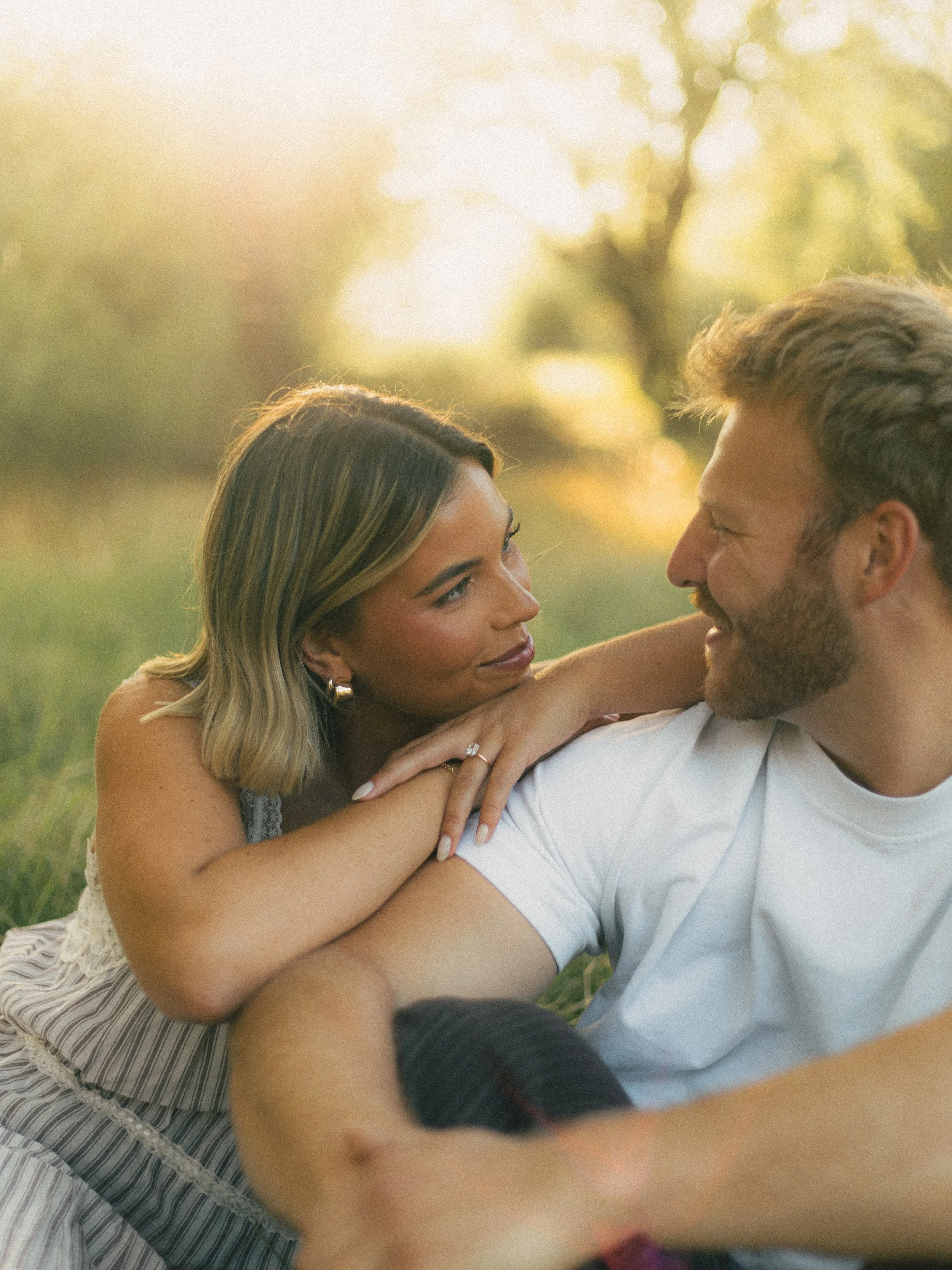 A woman and man sitting closely outside during sunset, gazing into each other's eyes with affectionate expressions.