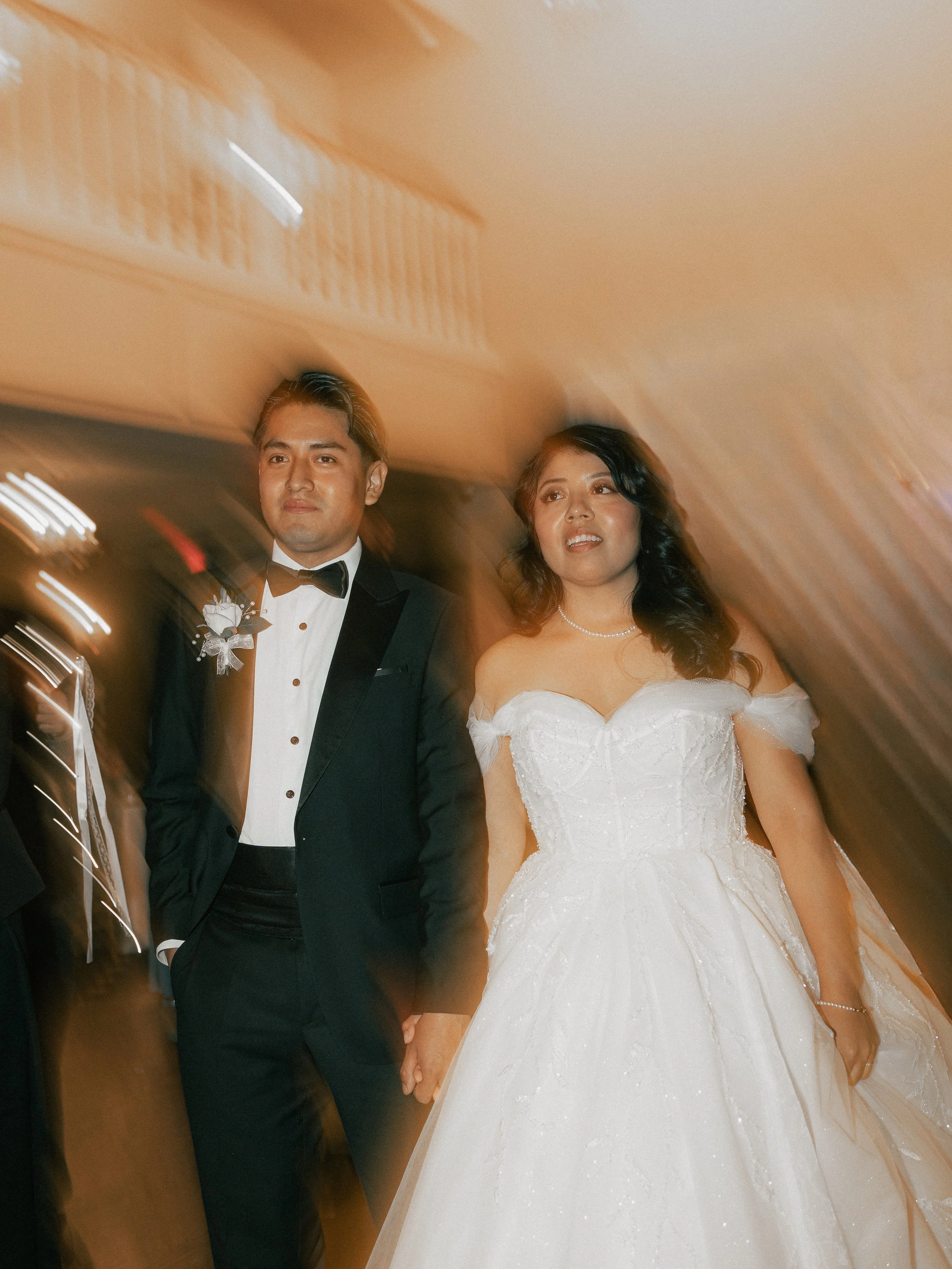 A bride and groom holding hands, dressed in wedding attire, standing in an elevator with warm lighting and wooden paneling.