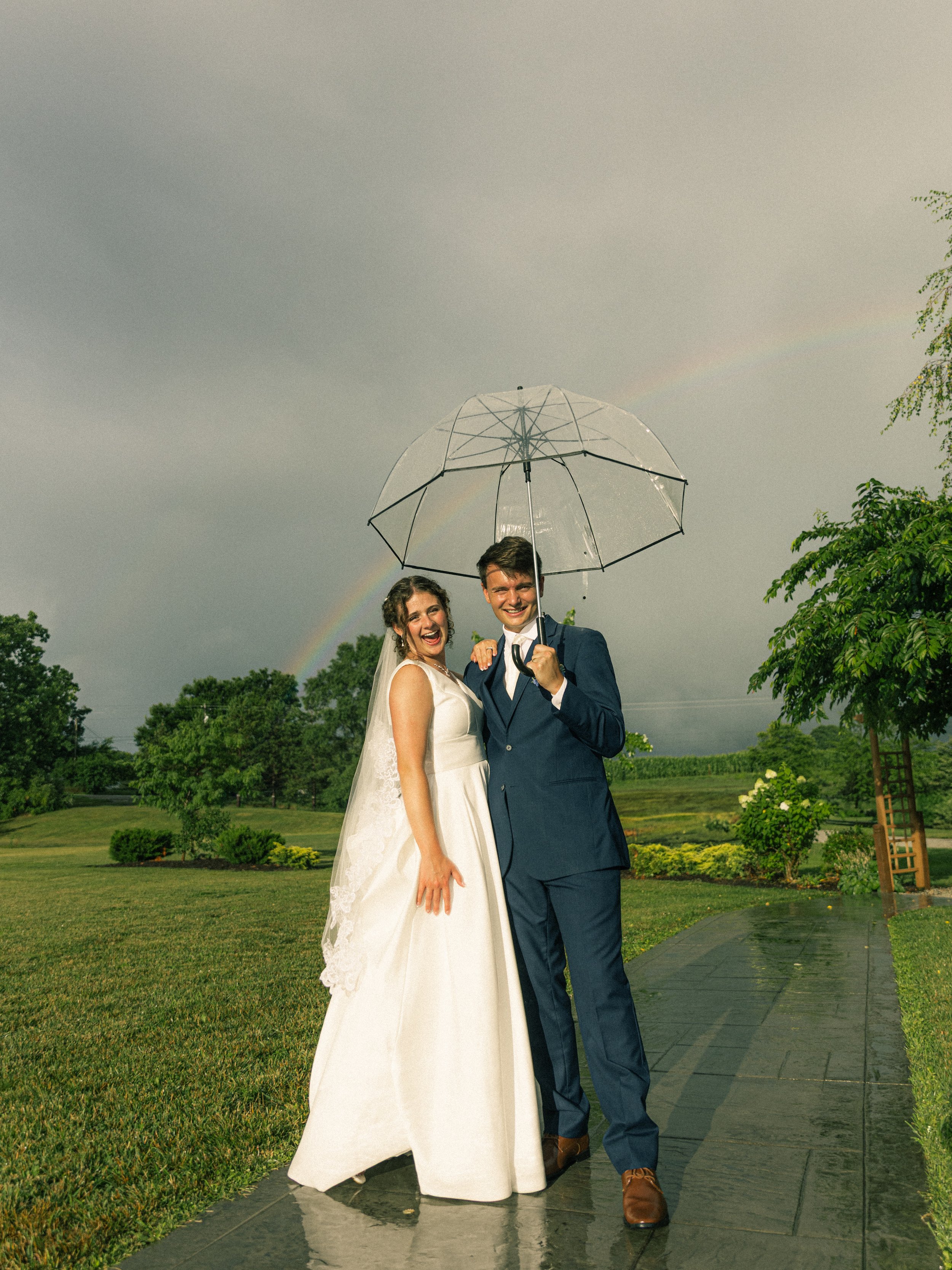 A bride and groom smiling under a transparency umbrella outdoors on a rainy day, with a rainbow visible in the background, and trees and a garden surrounding them.