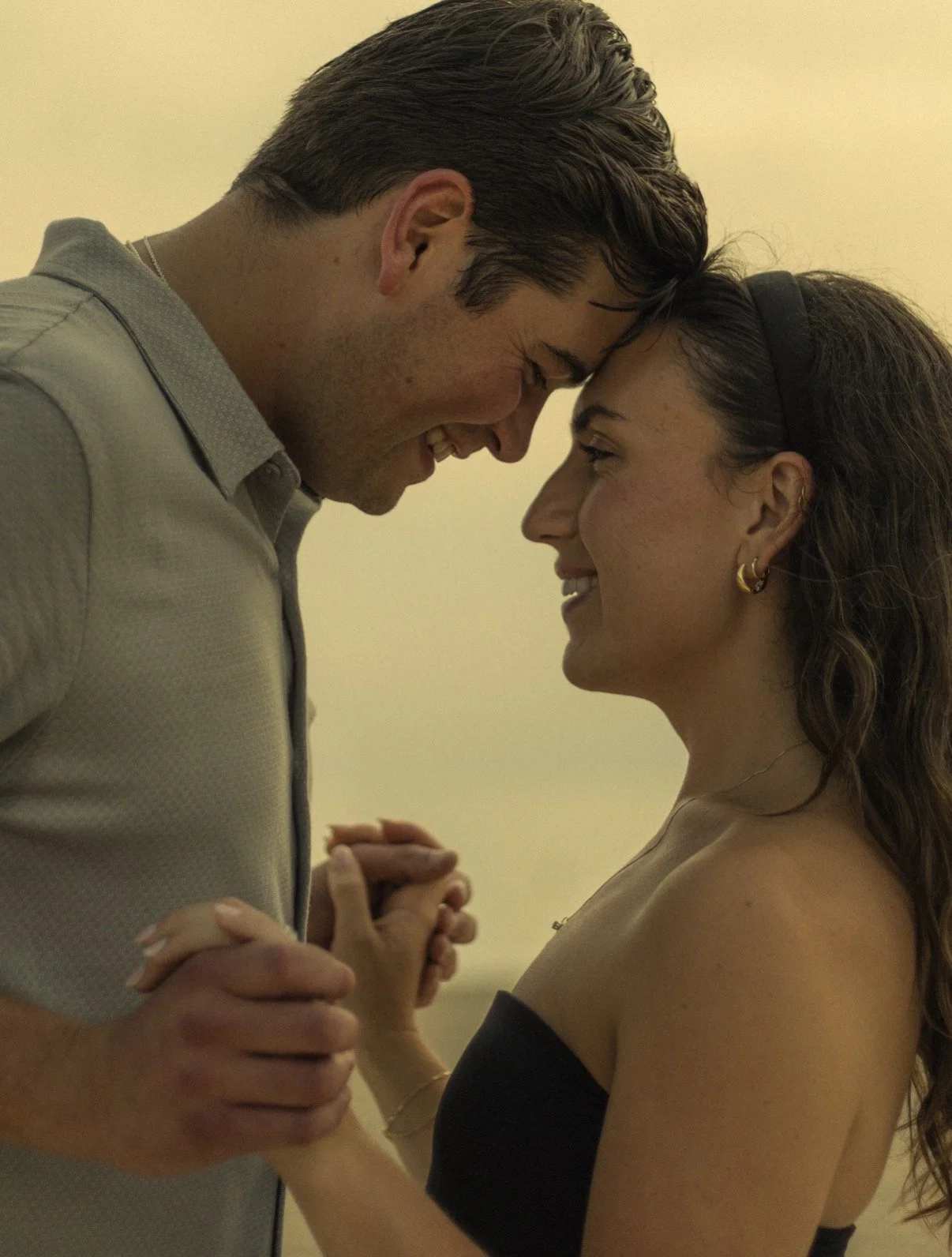 A man and a woman are dancing closely, with their foreheads touching and smiling at each other, holding hands, in a warm and intimate moment.