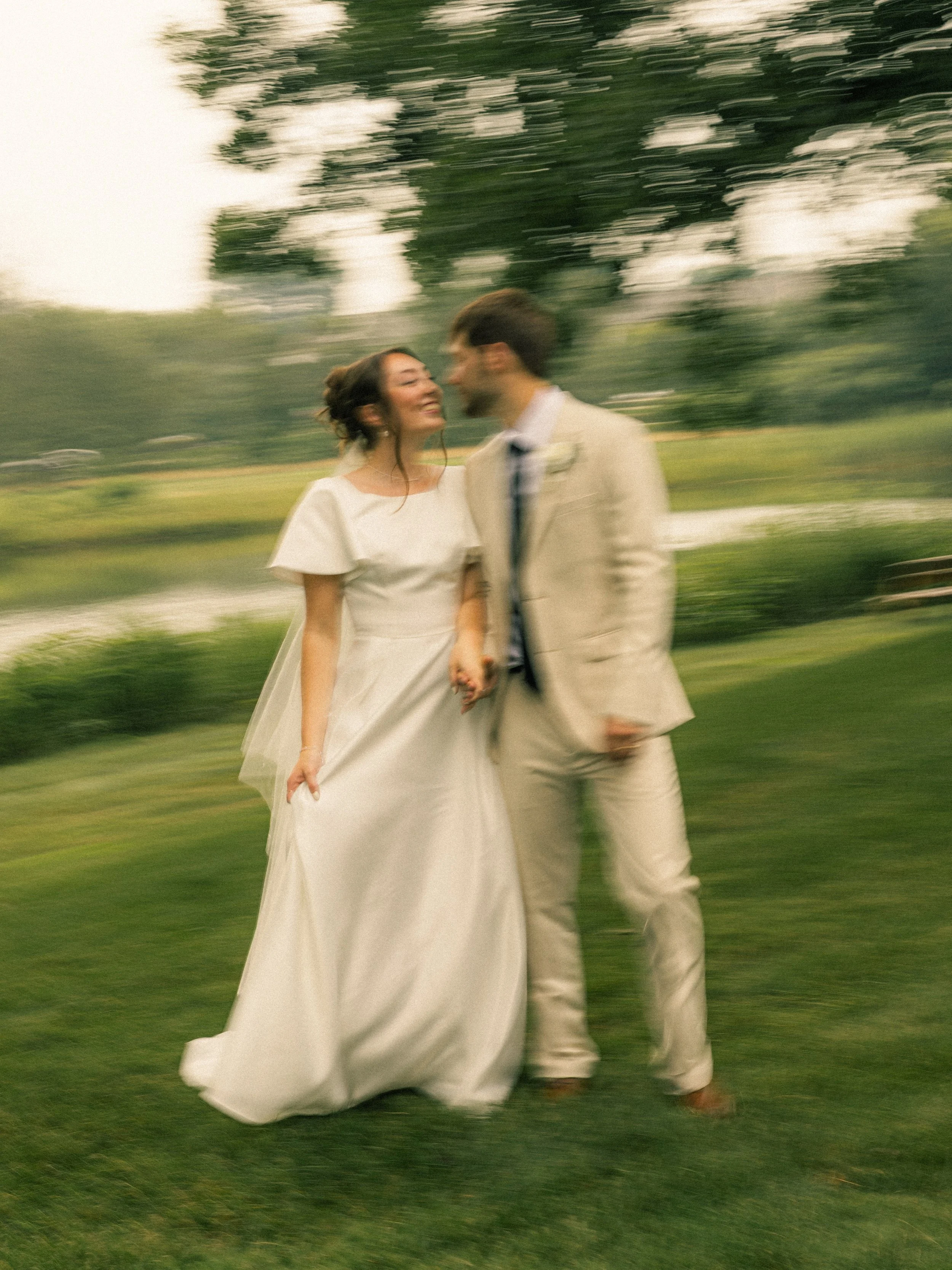 A couple in wedding attire holding hands and smiling at each other outdoors on a grassy area with trees in the background, photo appears to be intentionally blurred.