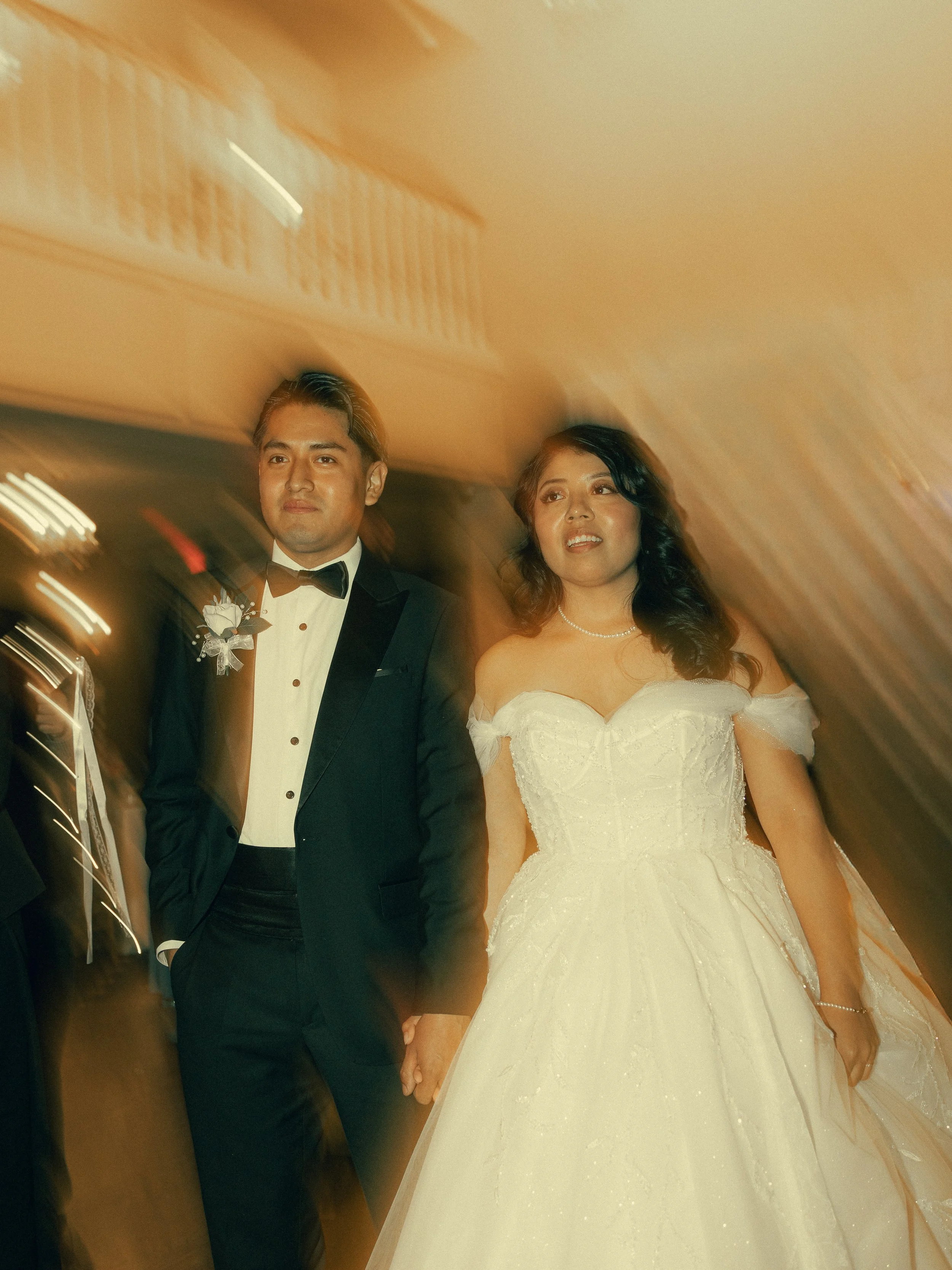 A newlywed couple holding hands in an elevator, dressed in formal wedding attire. The groom wears a black tuxedo with a bow tie, and the bride wears a white wedding gown with off-the-shoulder sleeves and pearl necklace. They are standing close togeth