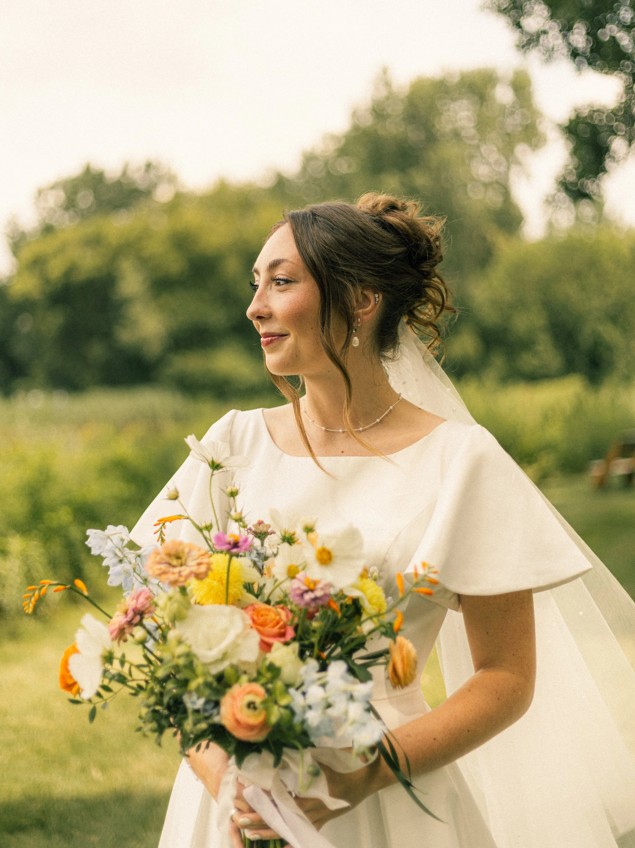 A bride in a white wedding dress holding a colorful bouquet of flowers outdoors with trees and greenery in the background