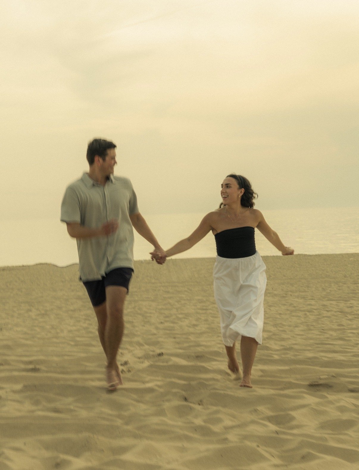 A man and woman holding hands and walking on a sandy beach, smiling at each other, during sunset.