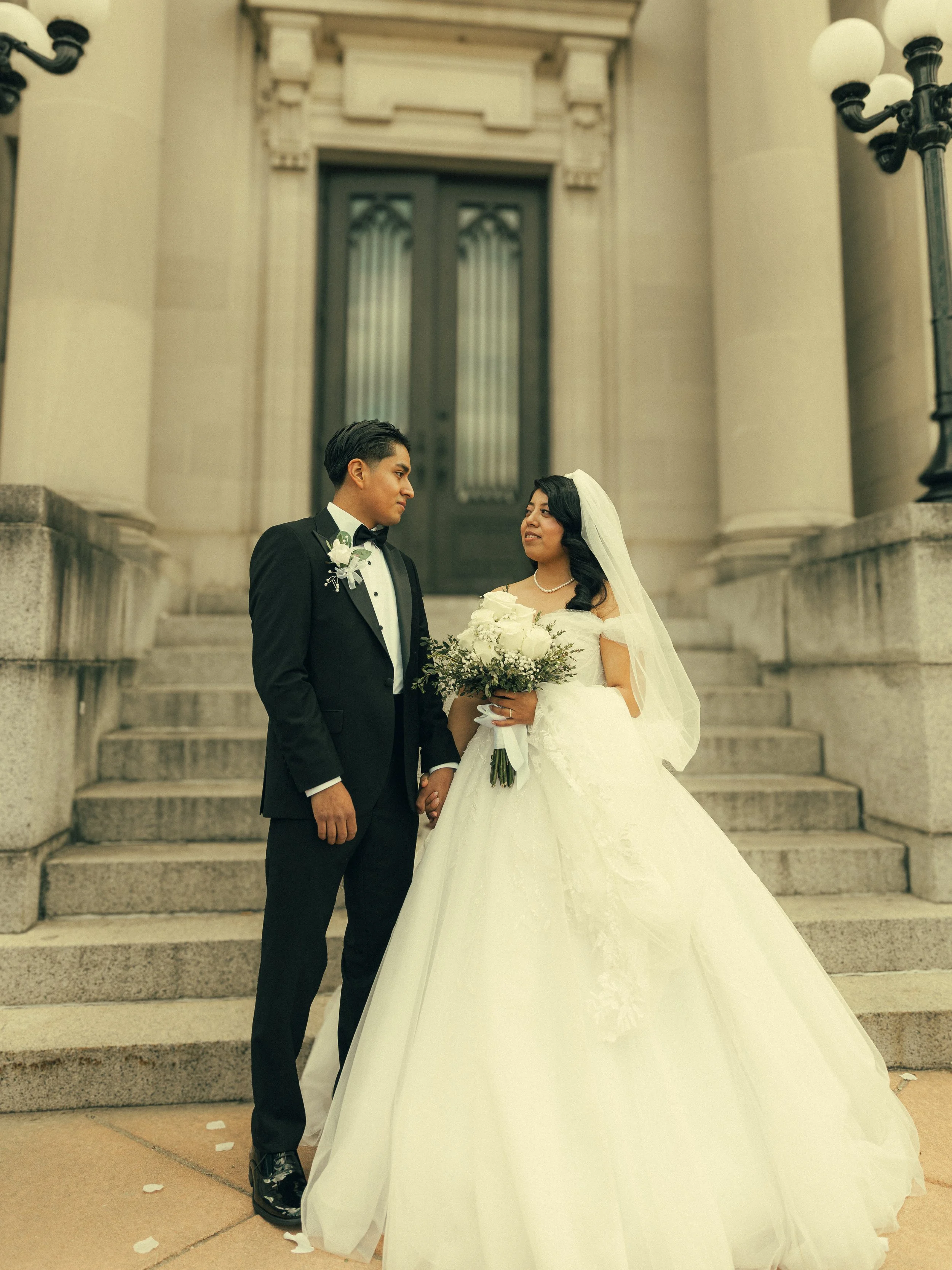 A bride and groom standing on the steps of a building, holding hands, with the bride holding a bouquet of white flowers. The groom is wearing a black tuxedo and the bride is in a white wedding gown with a veil.