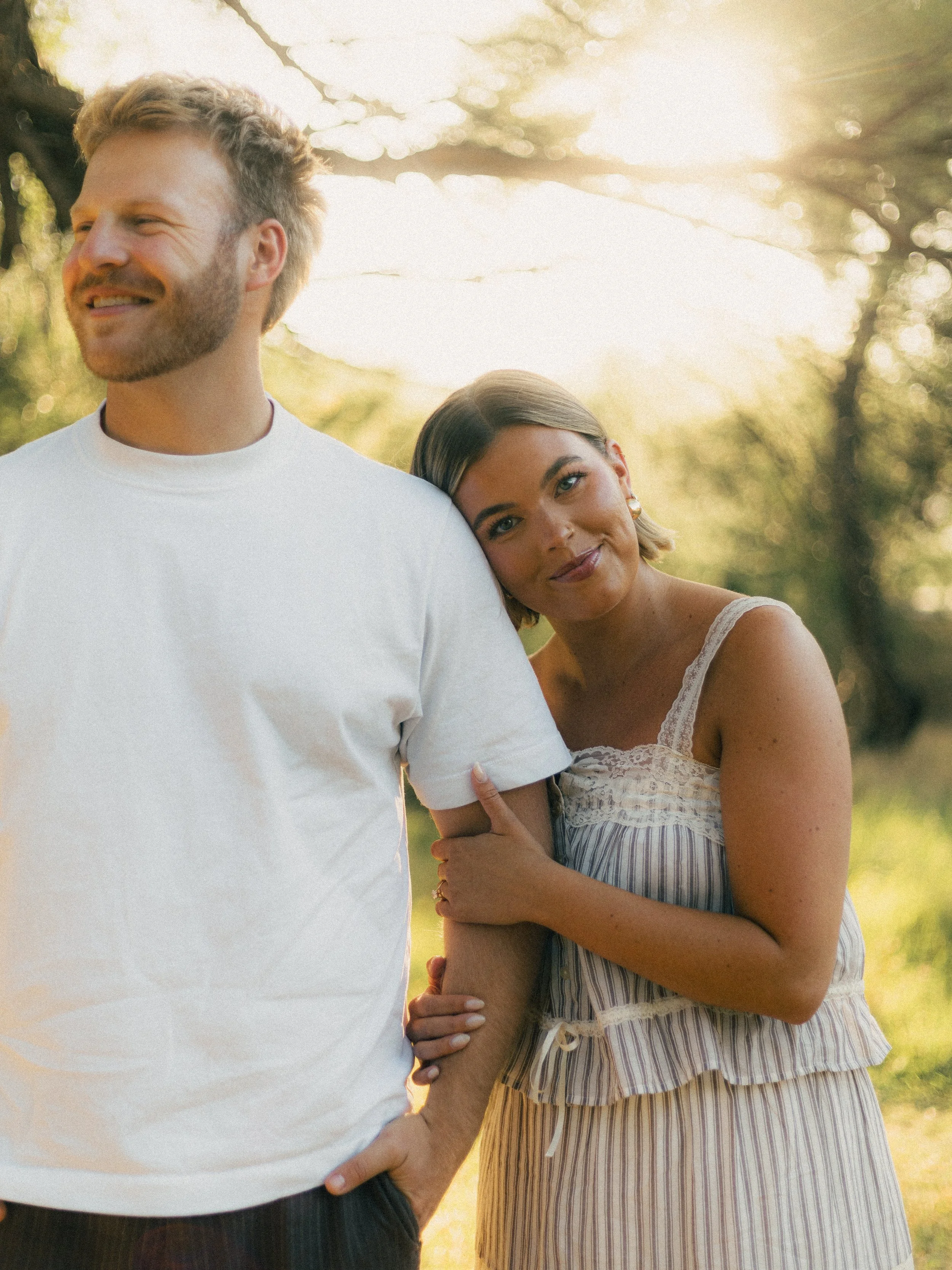A woman leaning her head on a man's shoulder, both smiling, standing outdoors during sunset with natural light and background trees.