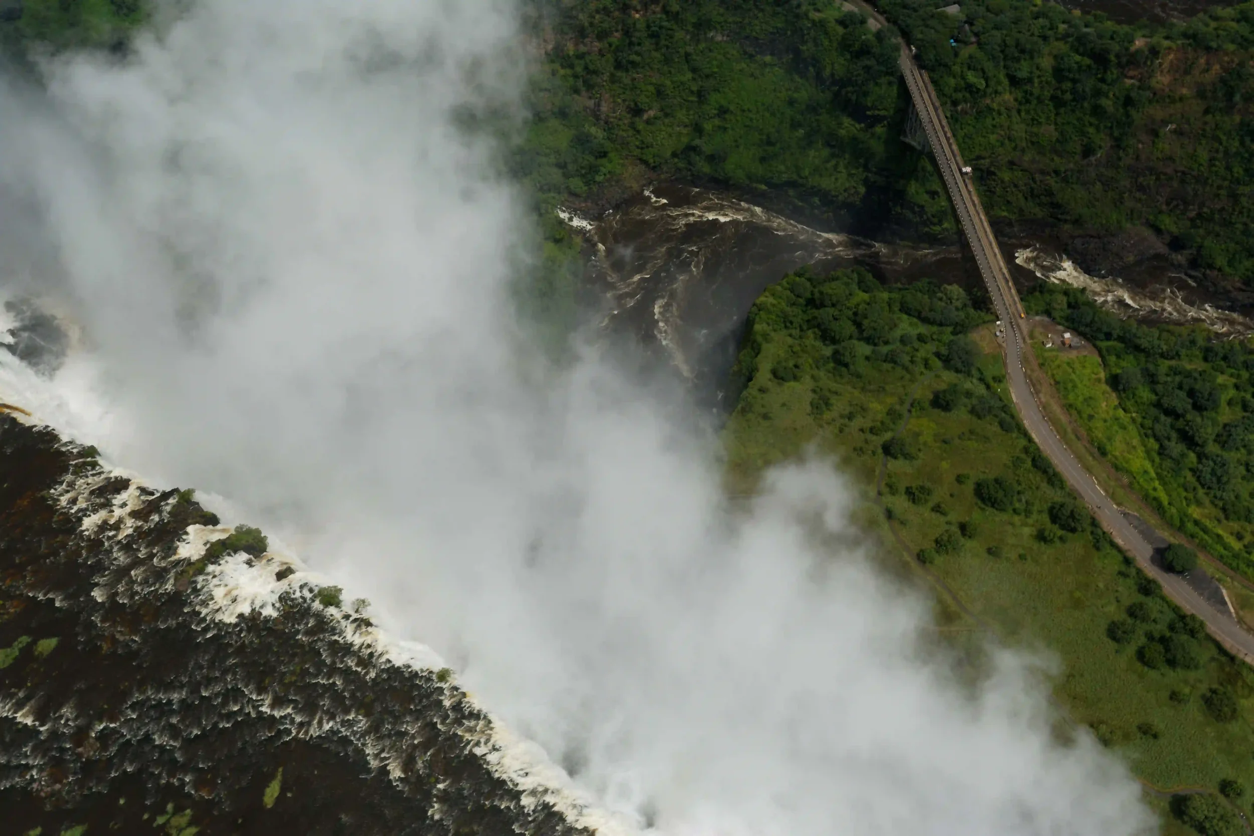 People walking on a narrow victoria falls suspension bridge with a metal railing and view of lush greenery below.