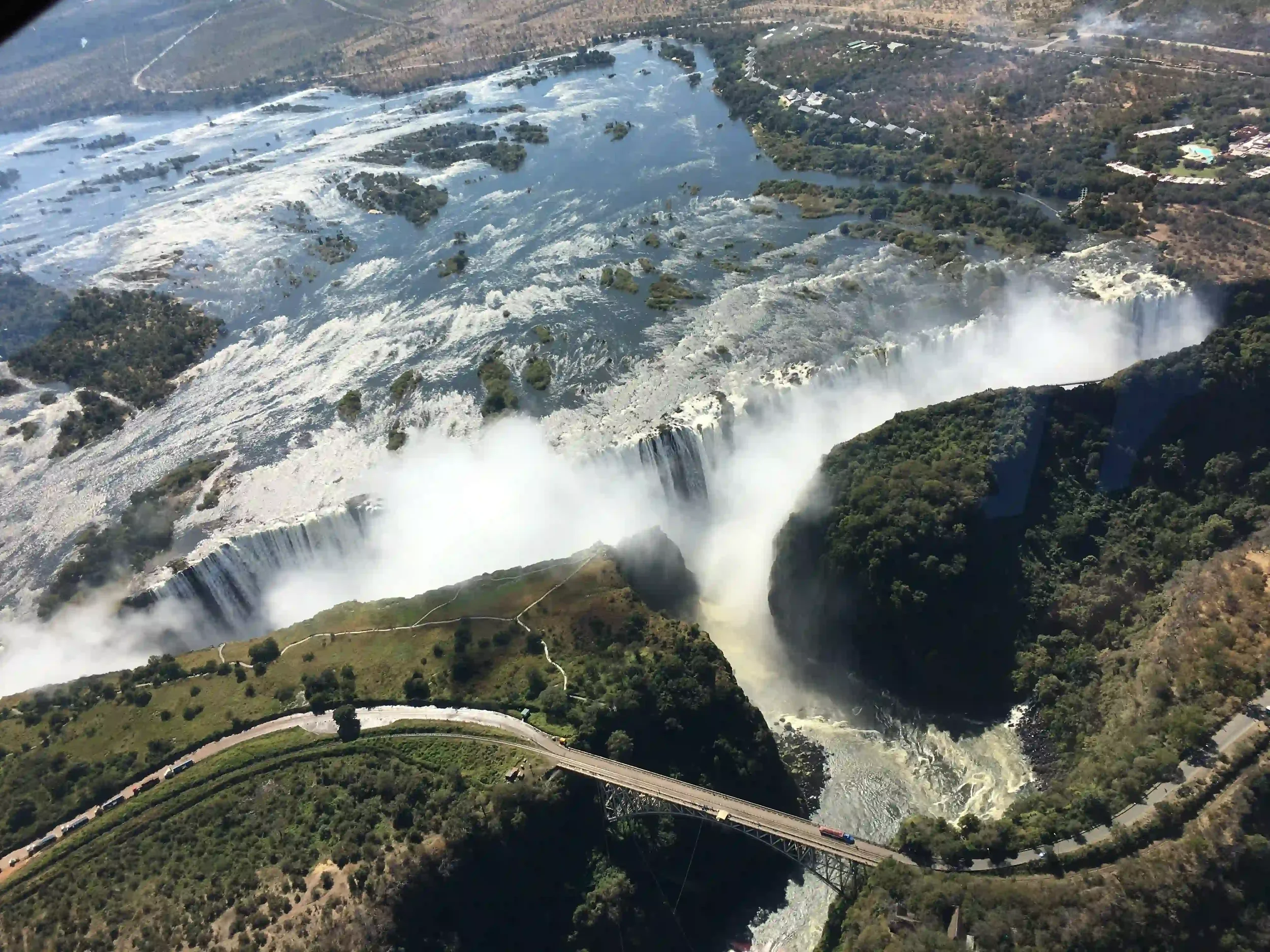 An aerial view of victoria Falls with a rainbow near the water spray, a bridge crossing below the falls, and lush green trees surrounding the area.