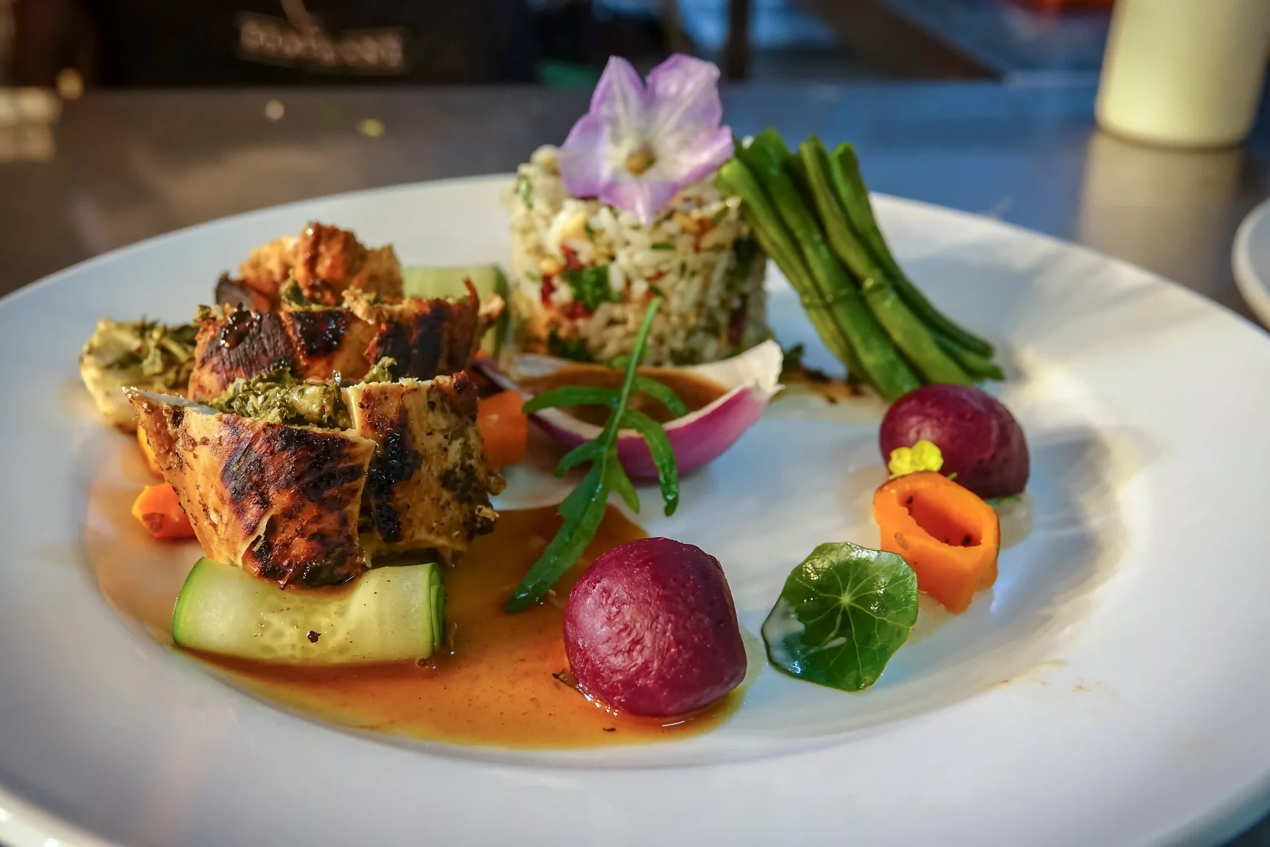 A plate of gourmet food featuring grilled meat, rice salad with herbs and vegetables, sliced cucumbers, radishes, and edible flowers for garnish.