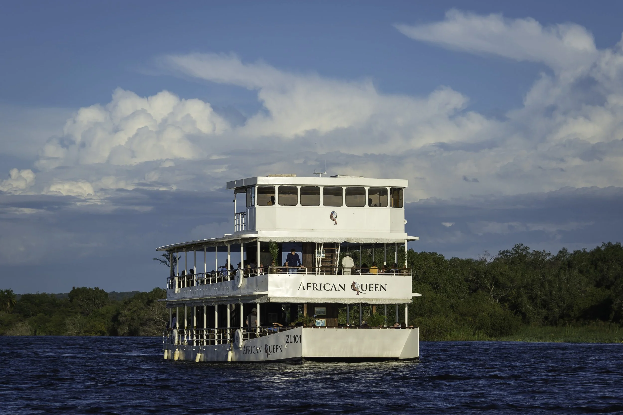 A white multi-deck boat named African Queen sailing on a body of water with green trees and a blue sky with scattered clouds in the background.