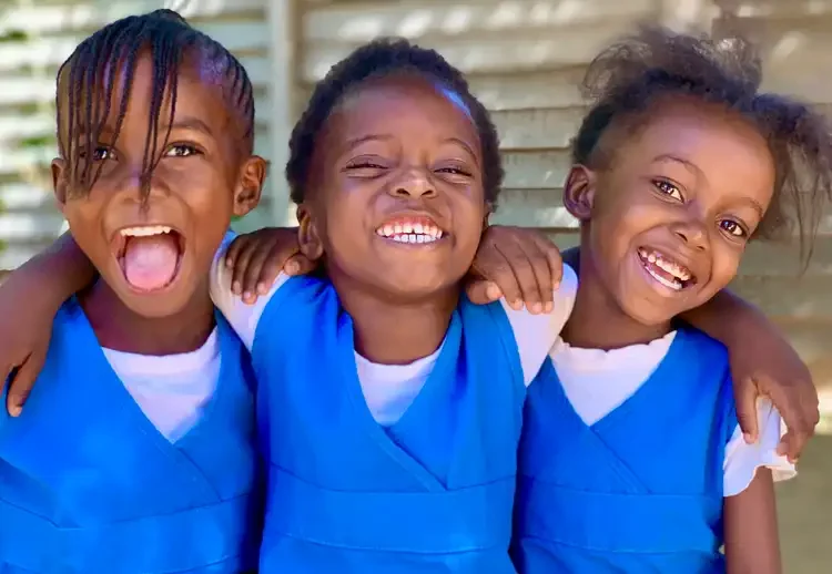 Three young girls with dark skin and natural hair smiling and hugging each other, wearing matching blue uniforms, in front of a wooden fence.