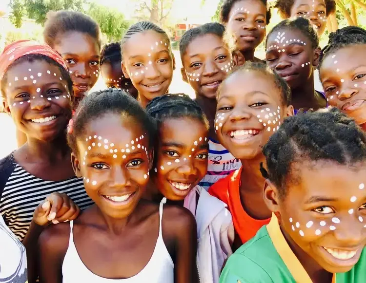 Group of smiling African girls with face paint, outdoors with trees in background.