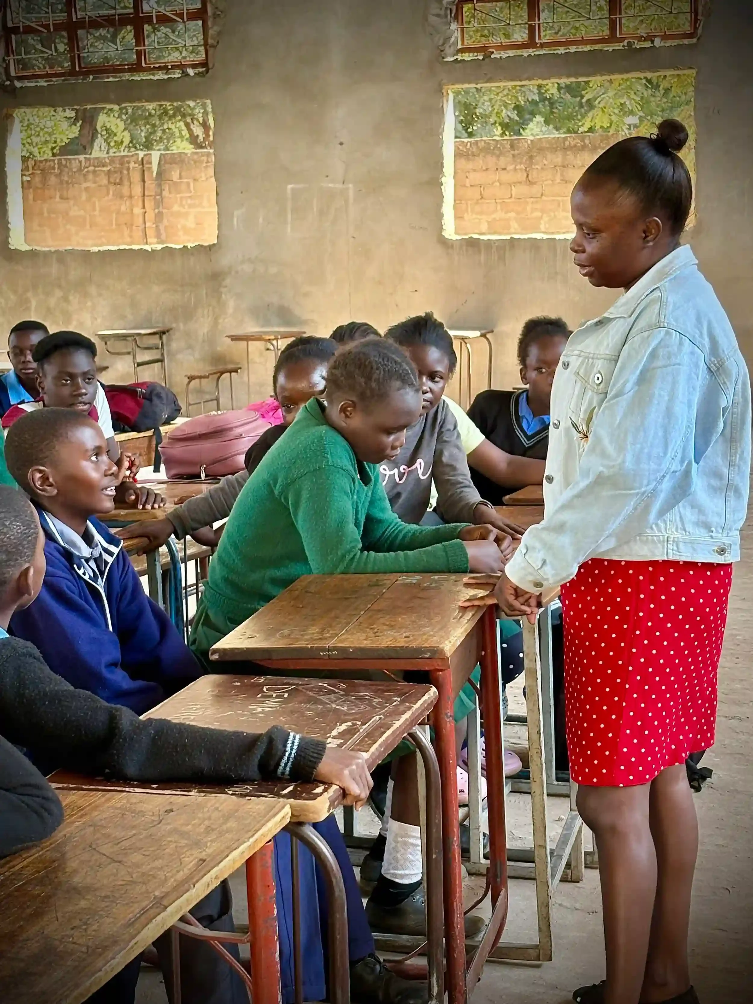 A classroom scene with students sitting at wooden desks listening to a teacher standing beside them. The classroom has unfinished walls with brick and concrete, and windows with bars showing green foliage outside.