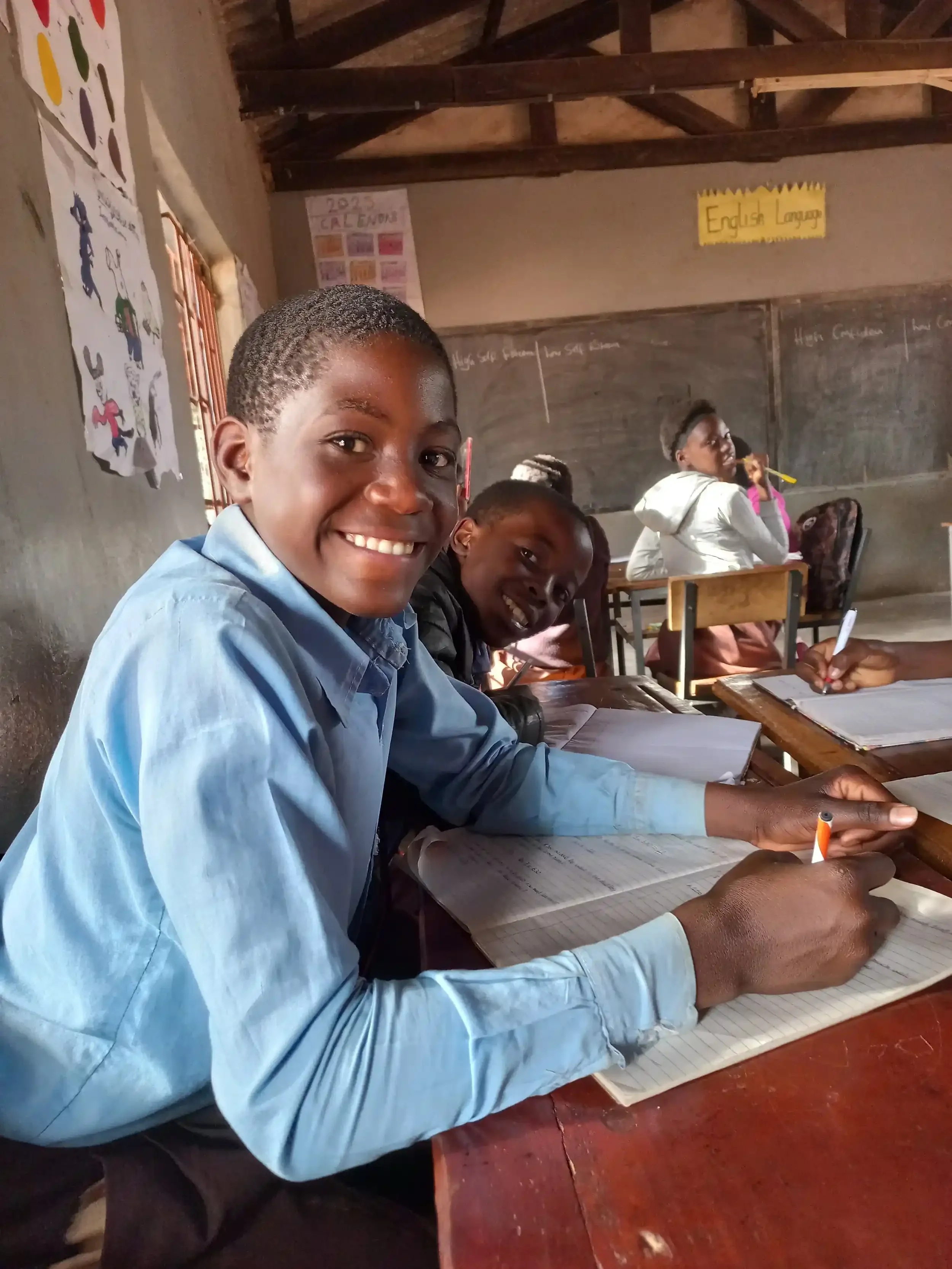 Students sitting in a classroom, smiling and taking notes with open notebooks and pens. The classroom has a chalkboard and educational posters on the wall.