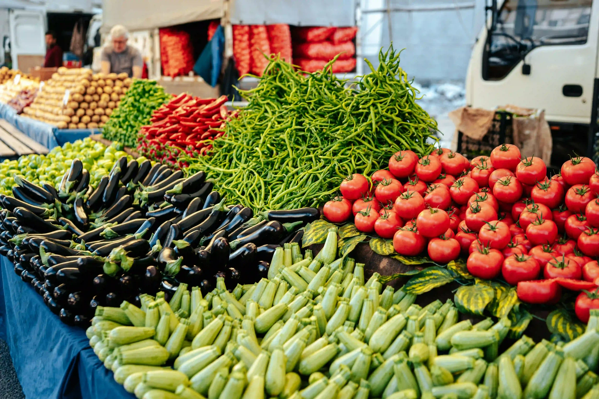 Display of fresh vegetables at an outdoor market, including tomatoes, green beans, zucchini, eggplants, and peppers.
