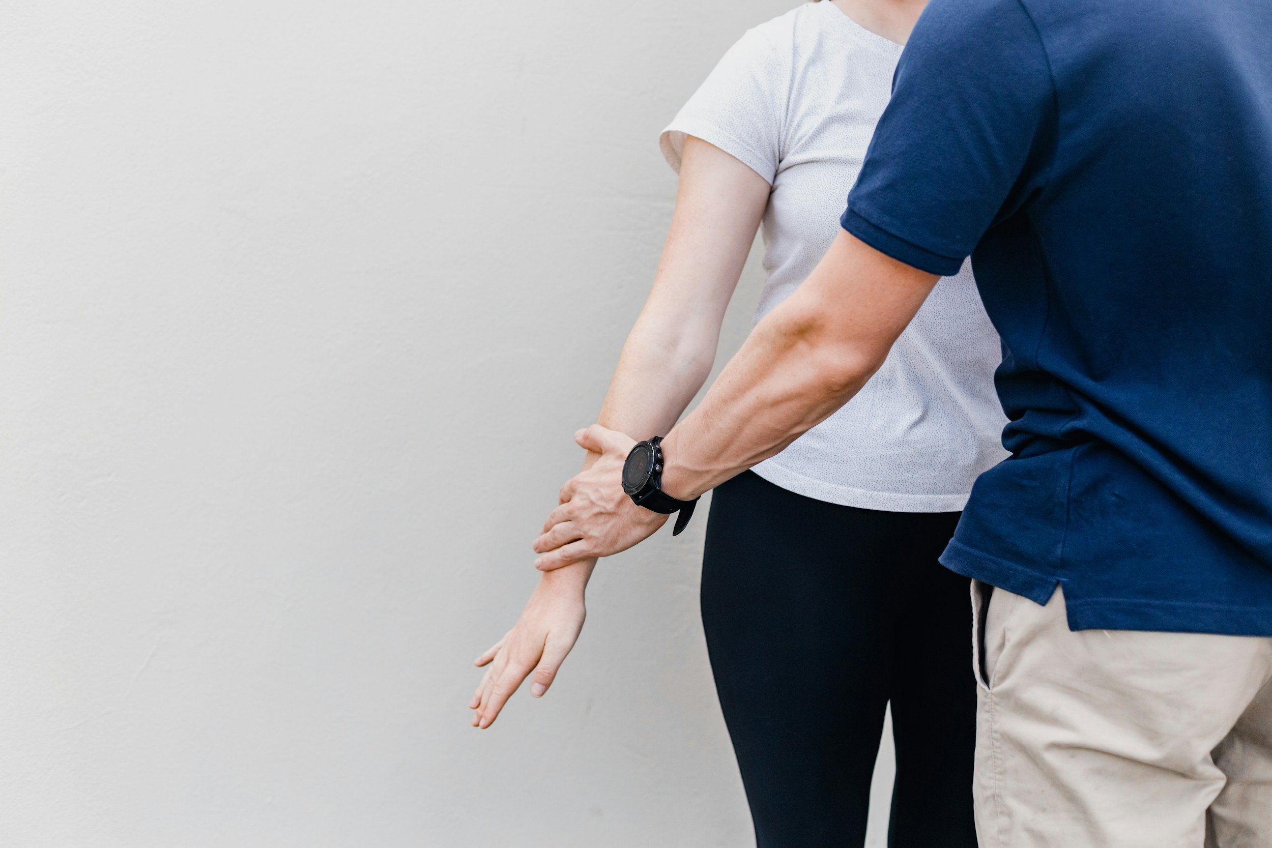 Two people are standing side by side against a plain white wall, with their arms extended downward. One person is wearing a white shirt and the other is wearing a blue shirt, and both are wearing watches on their left wrists.