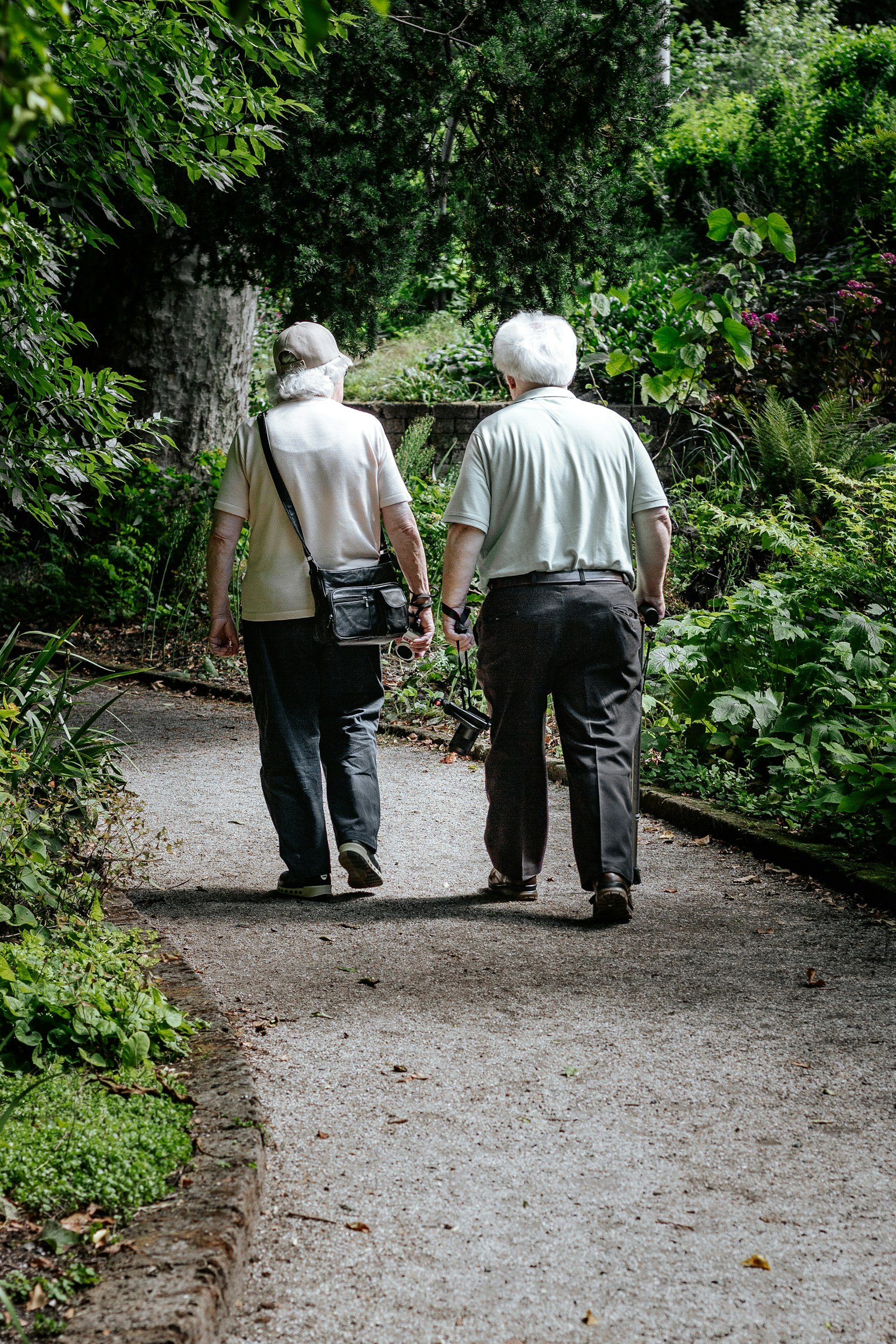 Two elderly people with gray hair, wearing beige clothing, walking together on a forest trail surrounded by green foliage.