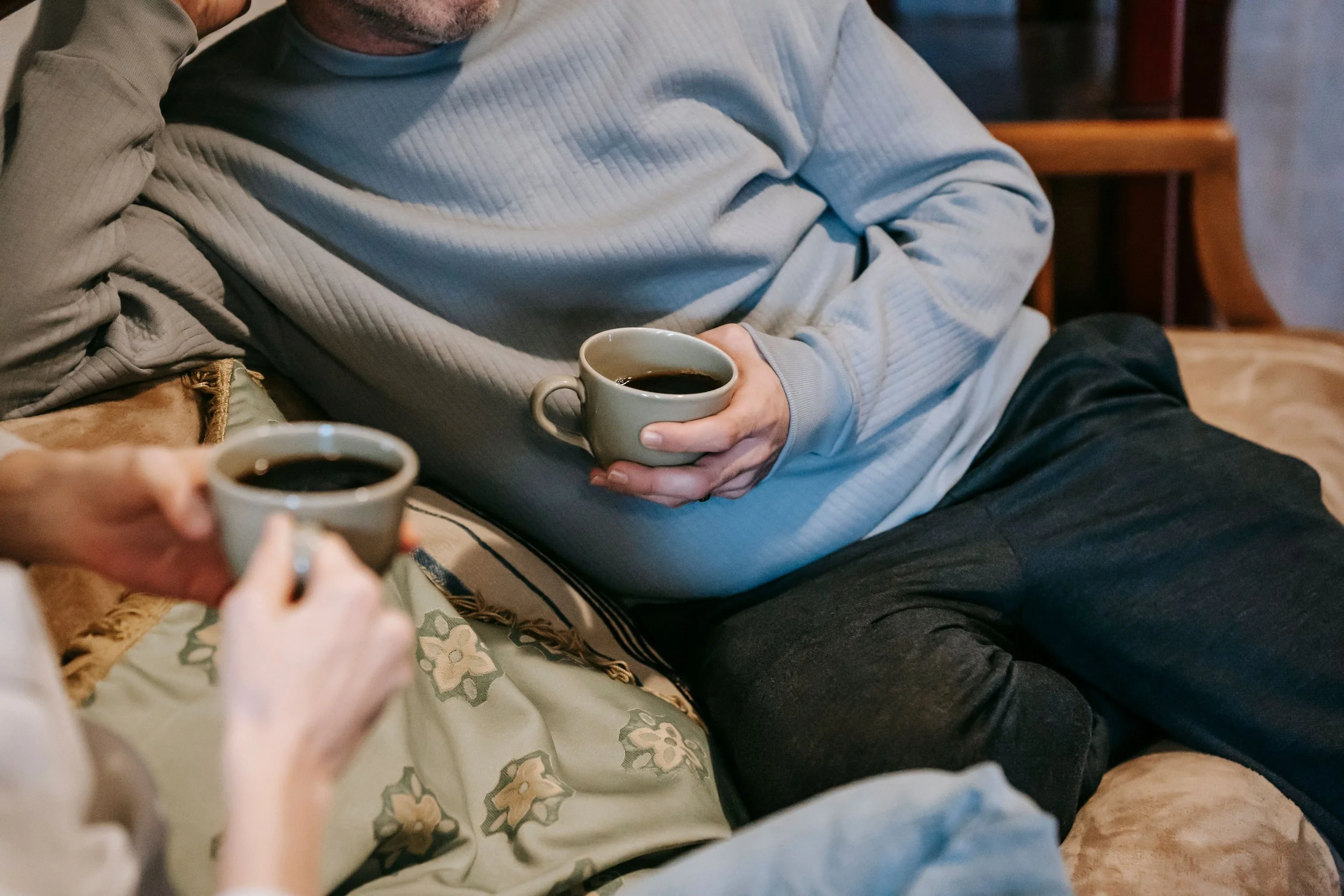 Two people sitting on a couch or chair, holding cups of coffee or tea. One person is wearing a light blue or gray long-sleeve shirt, and the other has a white or beige top. The setting appears cozy and homely.
