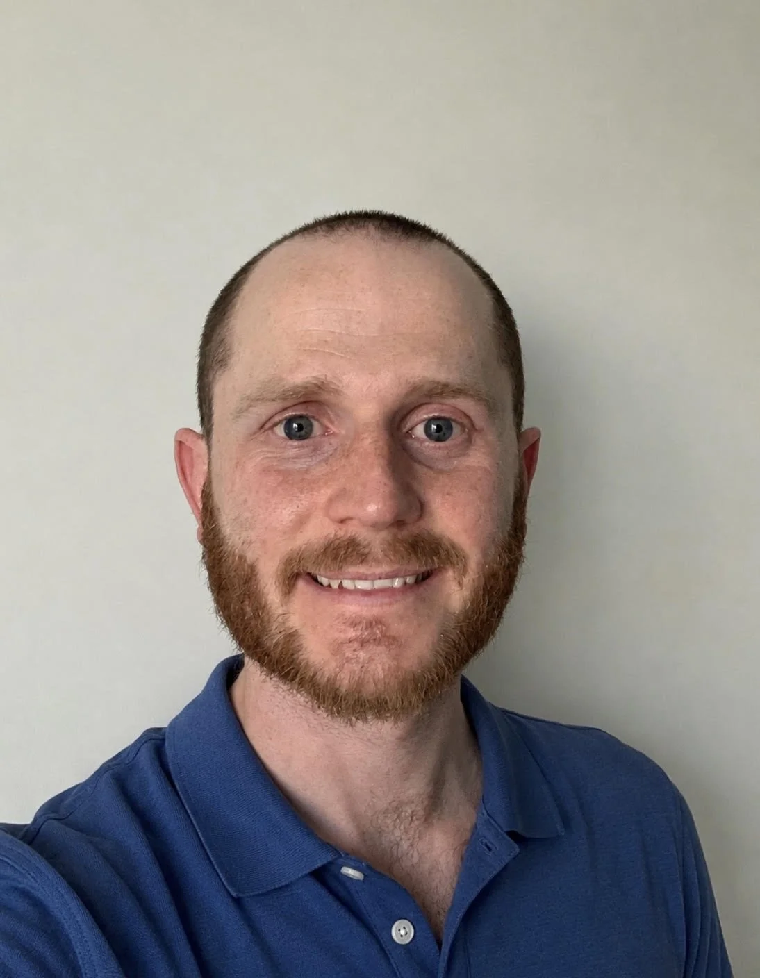 A man with short hair and a beard smiling, wearing a blue collared shirt, standing against a plain light-colored wall.