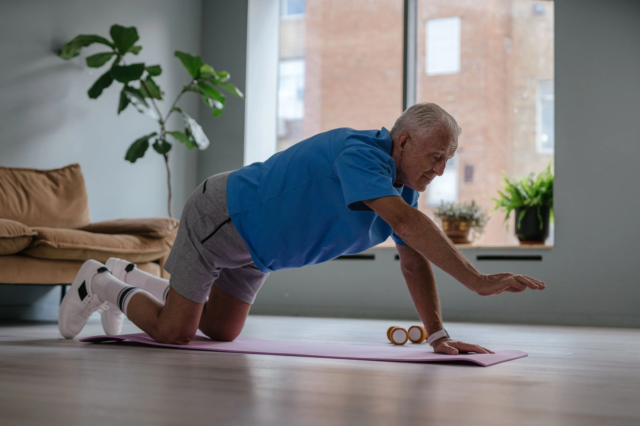 An elderly man with white hair doing a plank or superman exercise on a pink yoga mat in a bright room with large windows, plants, and a brown sofa.