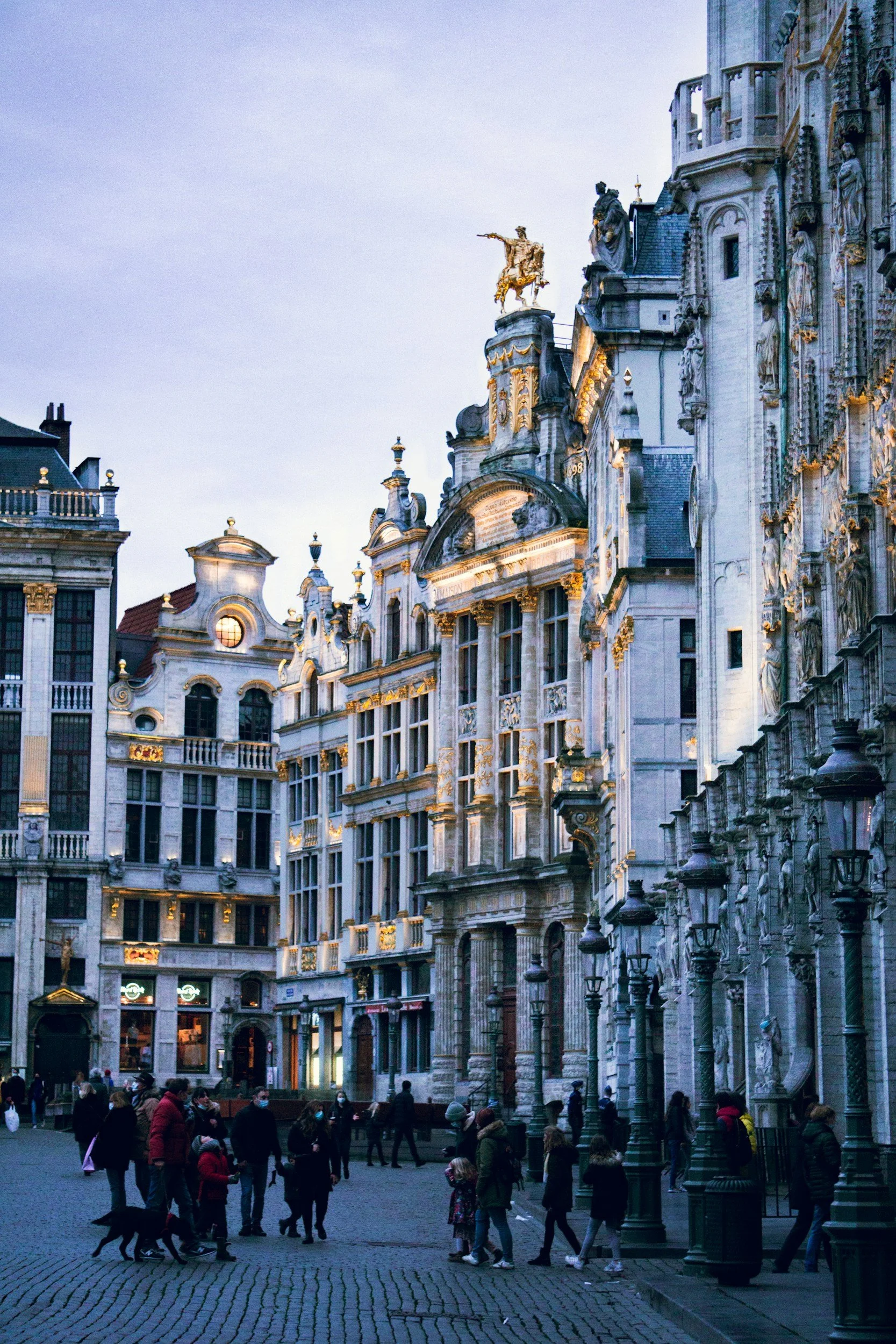 City square in Europe with historic architecture, people walking, and street lamps at dusk.