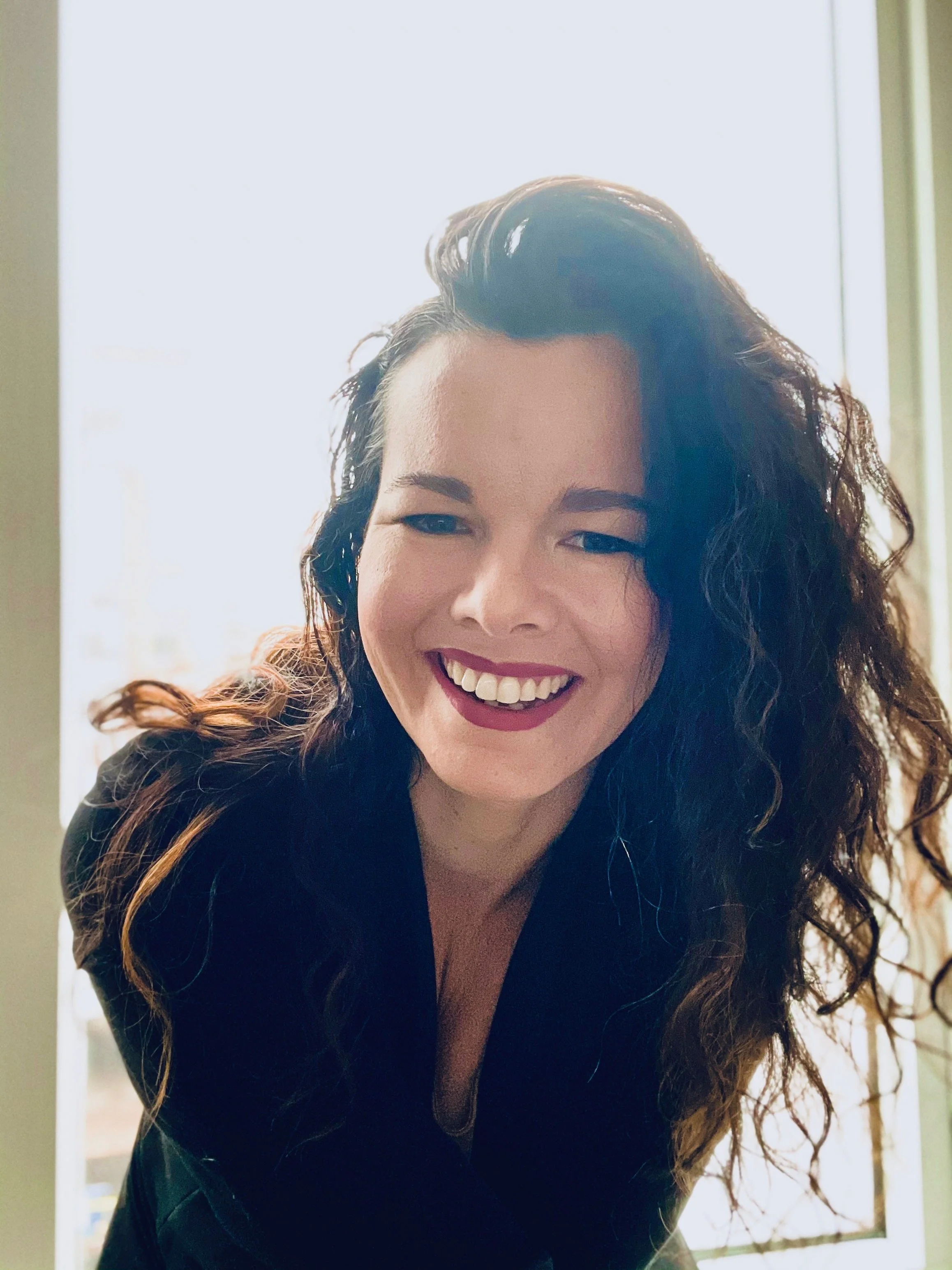 A woman smiling and leaning towards the camera, with dark, curly hair and wearing a black top, standing near a window with light behind her.