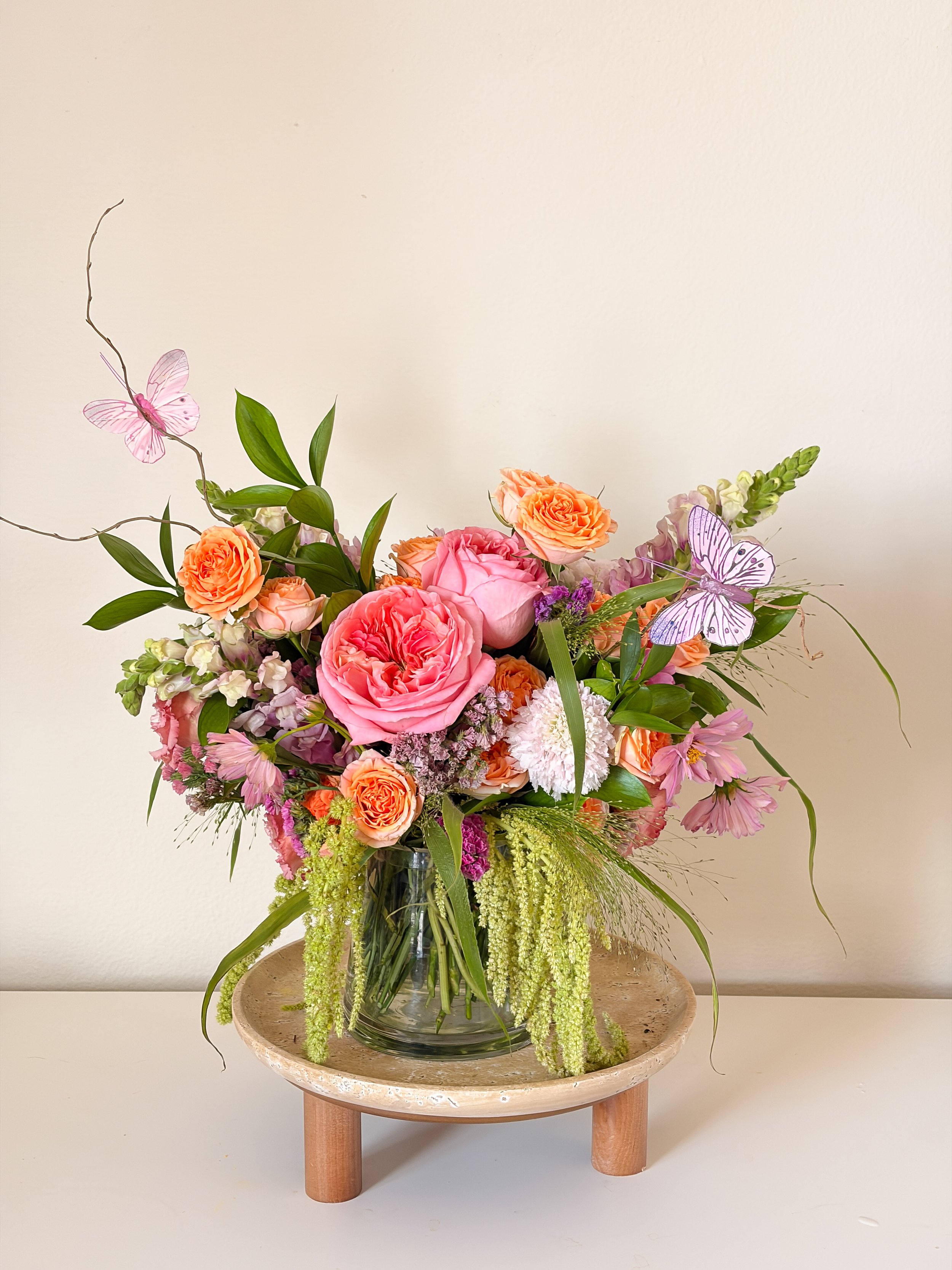 A bouquet of pink and orange flowers with greenery and two purple butterflies in a clear glass vase on a round wooden table against a plain white background.