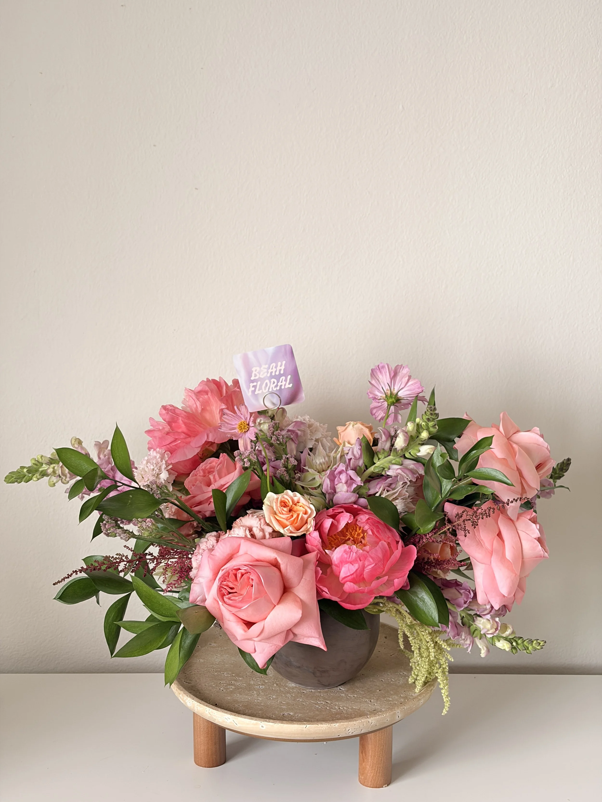 A colorful bouquet of pink and purple flowers in a brown vase on a small wooden table against a plain white wall.