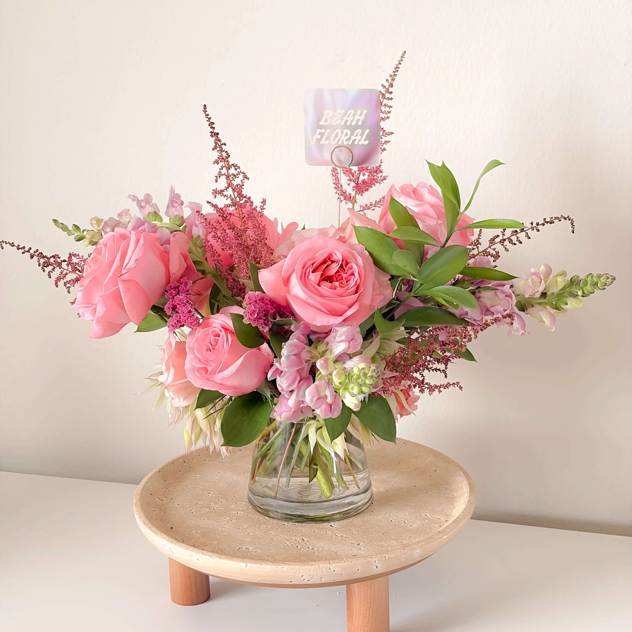 A floral arrangement in a clear glass vase with pink roses, purple flowers, and greenery, placed on a round beige marble table with wooden legs.
