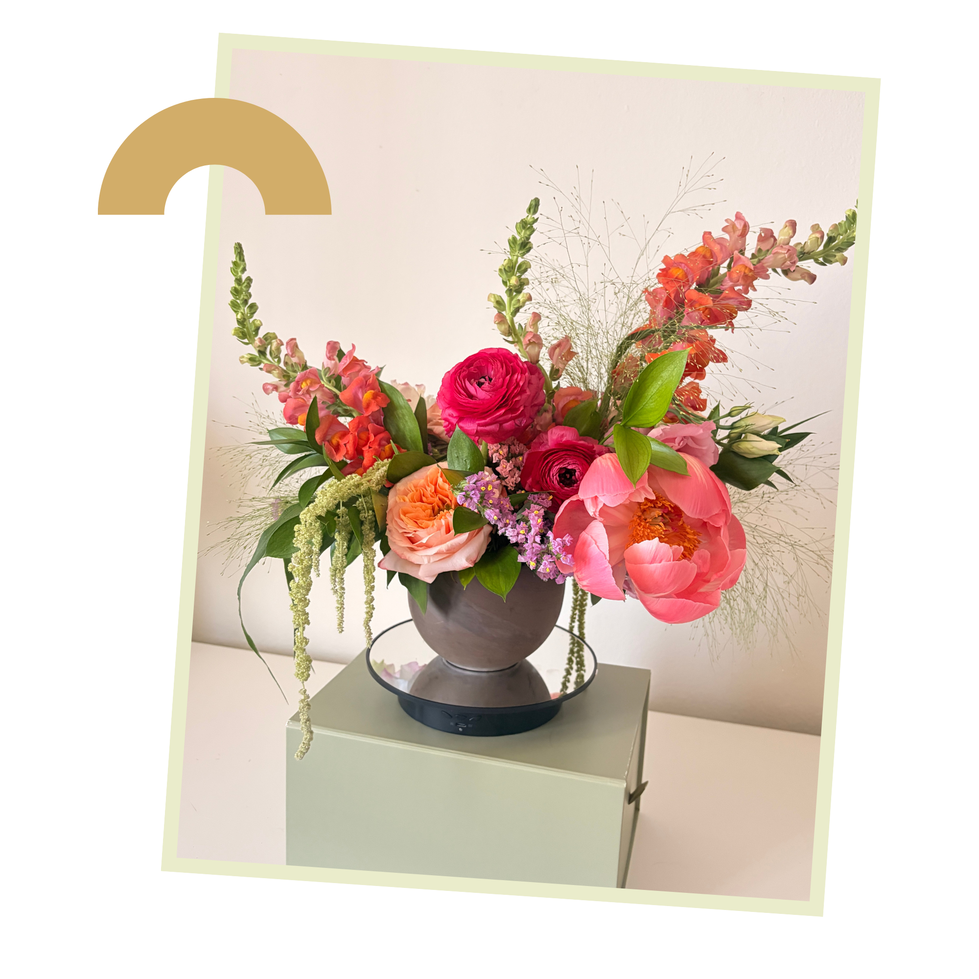 A colorful bouquet of flowers including pink and red roses, orange snapdragons, purple statice, and greenery in a dark gray vase, placed on a white surface against a white background.