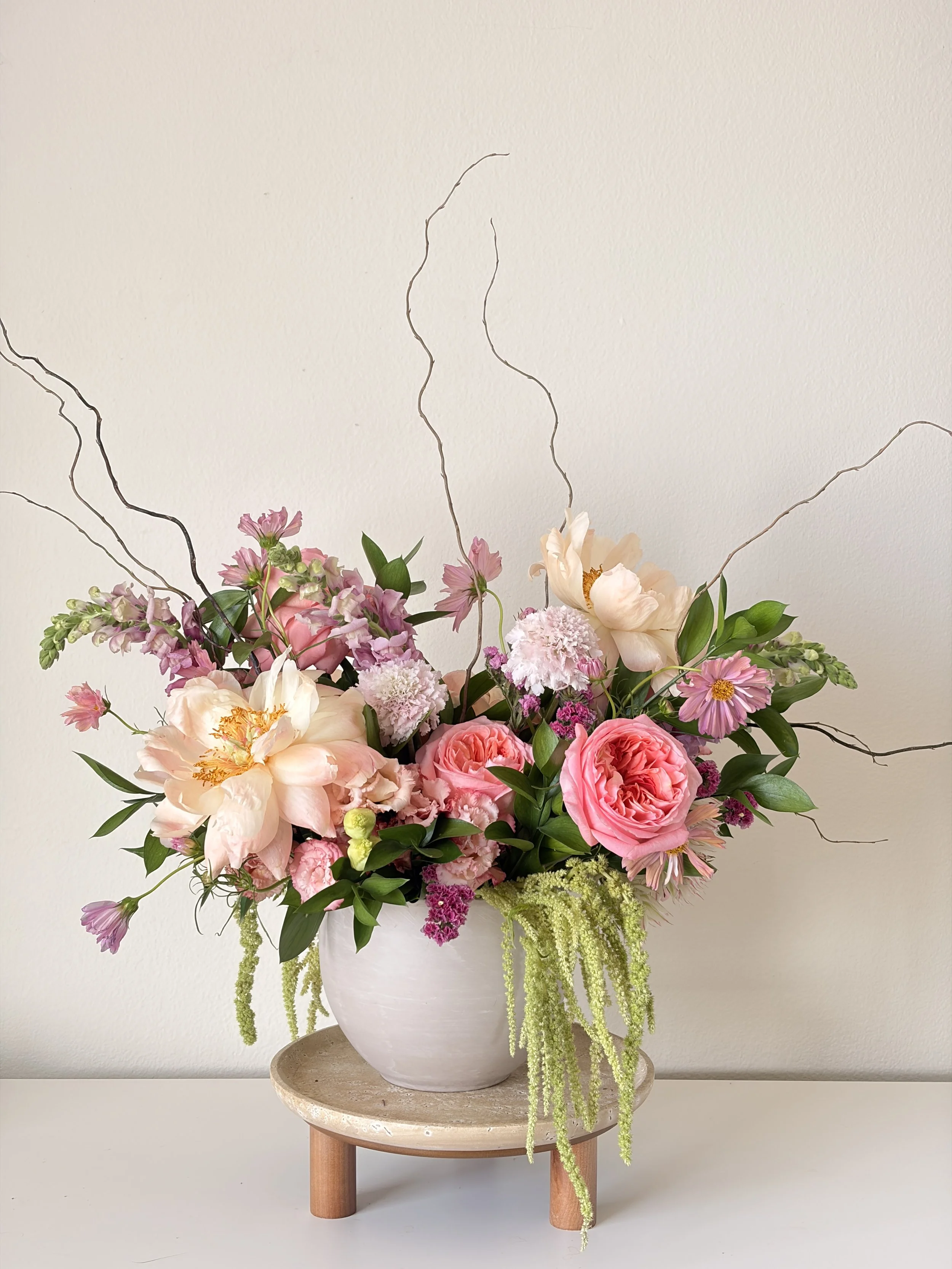 A floral arrangement in a white vase on a small wooden table against a white wall, featuring pink, peach, white flowers, and long green hanging plants.