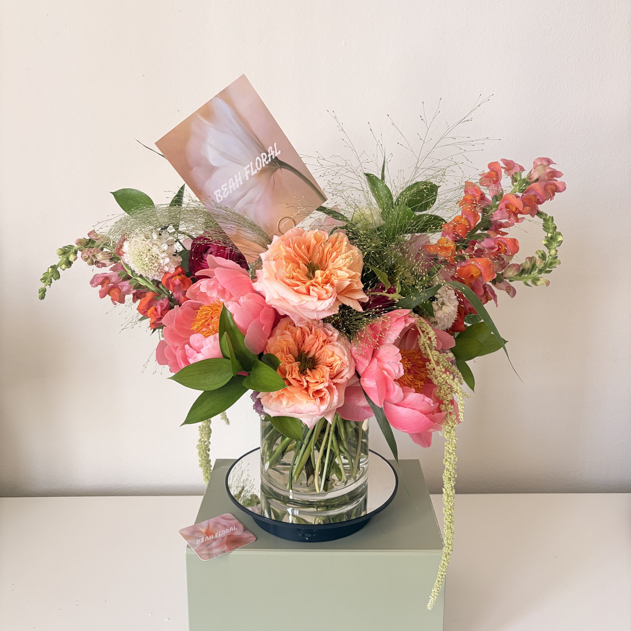 A floral arrangement in a glass vase includes pink, peach, and red flowers, with some green leaves and wispy greenery, with a small card reading 'BEAH FLORAL' placed in the bouquet.