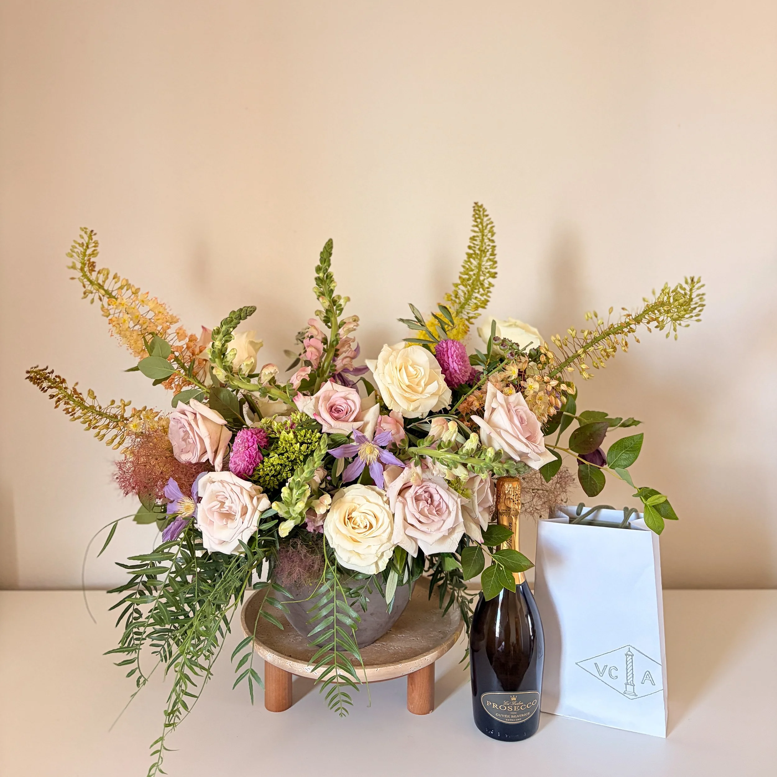 A arrangement of various flowers including white roses, pink roses, purple flowers, green leaves, and tall yellow and green sprigs in a large brown pot, with a bottle of Prosecco nearby and a white box with a label that reads 'VCΑ'.