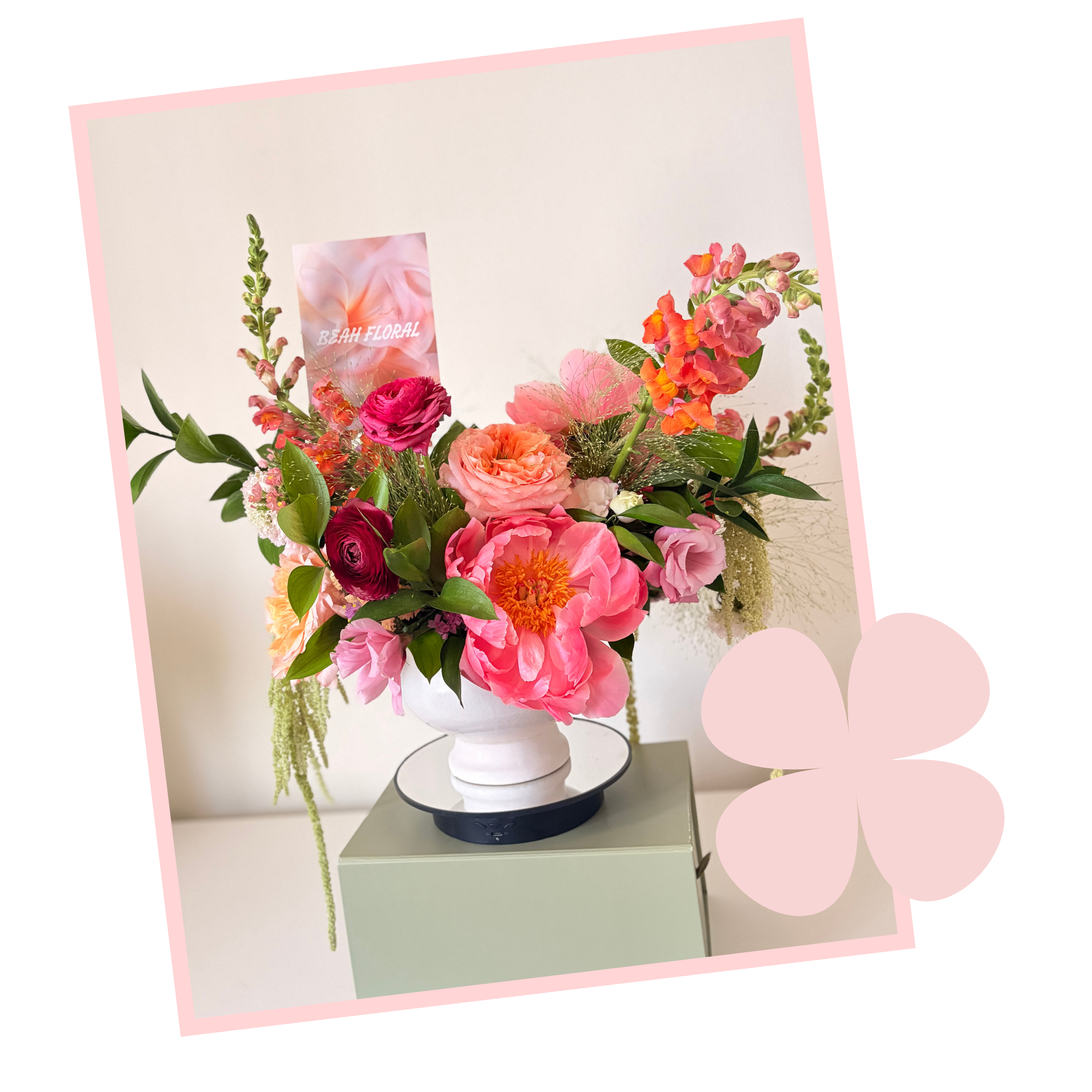 A floral arrangement in a white vase with pink roses, orange snapdragons, and greenery, placed on a small table with a digital speaker beneath it, against a light-colored background.