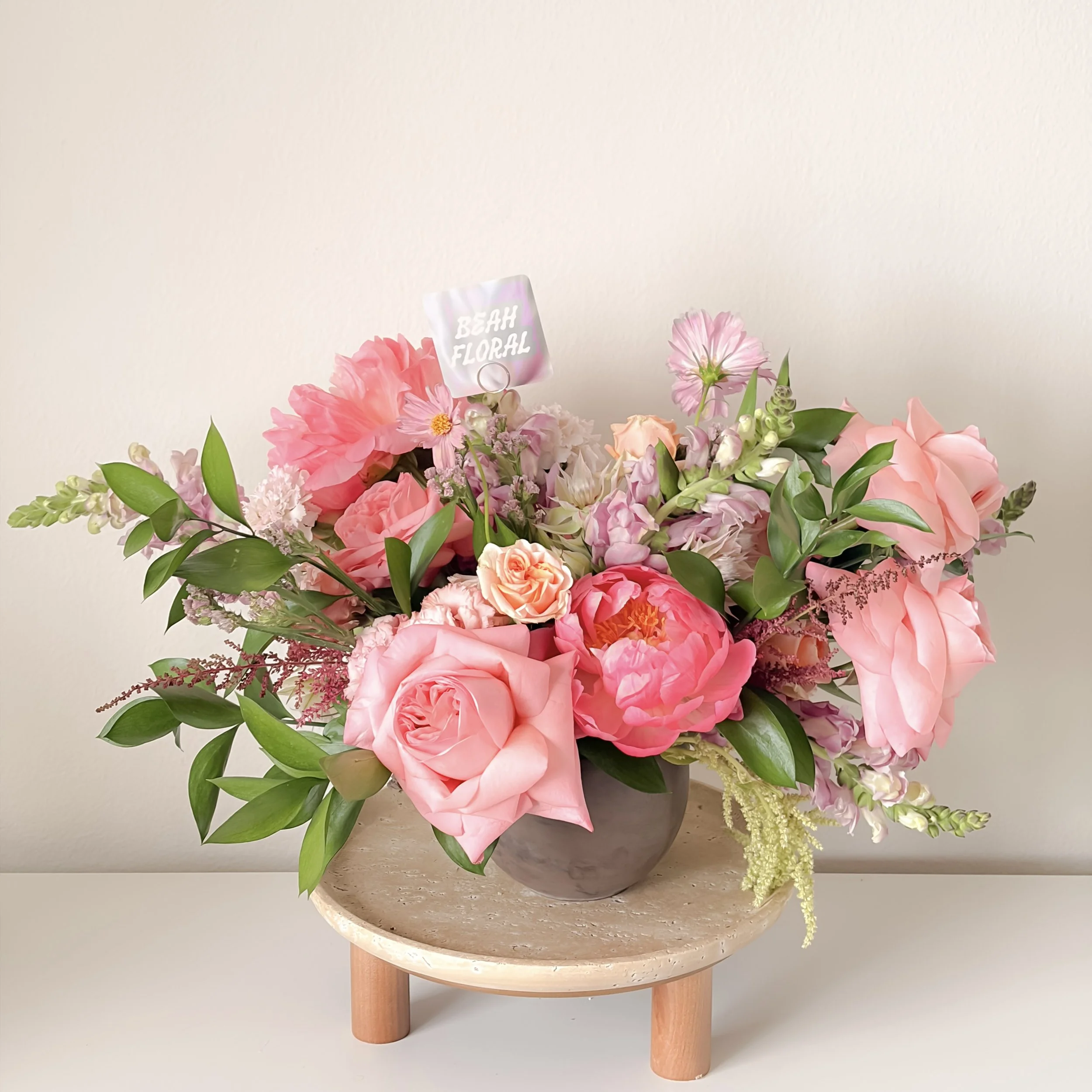 A large flower arrangement in a round ceramic pot with pink, peach, and white flowers and greenery, placed on a light-colored table against a plain wall, with a small tag that reads 'BEAH FLORAL'.