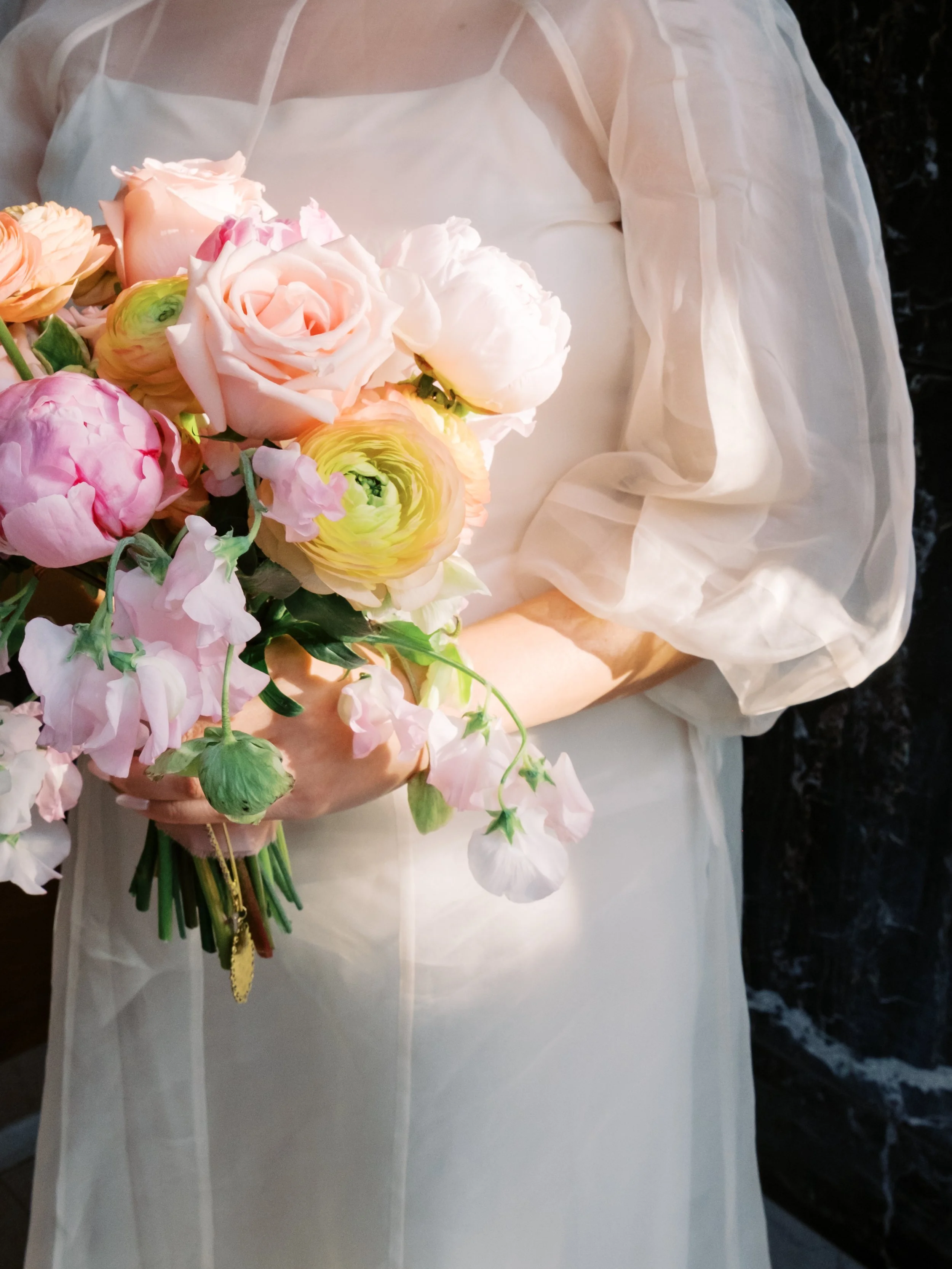 Close-up of a person holding a bouquet of pink, cream, and yellow flowers, wearing a white dress with sheer long sleeves.